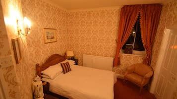 Un dormitorio con cama, una silla, un radiador, una ventana con cortinas y una lámpara fijada a la pared.