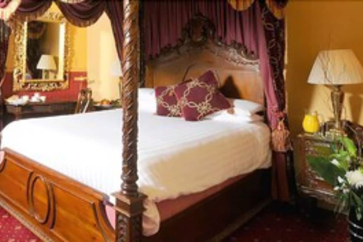 A four-poster bed with a canopy, a bedside table with a lamp, and a view of the outdoors through a window.