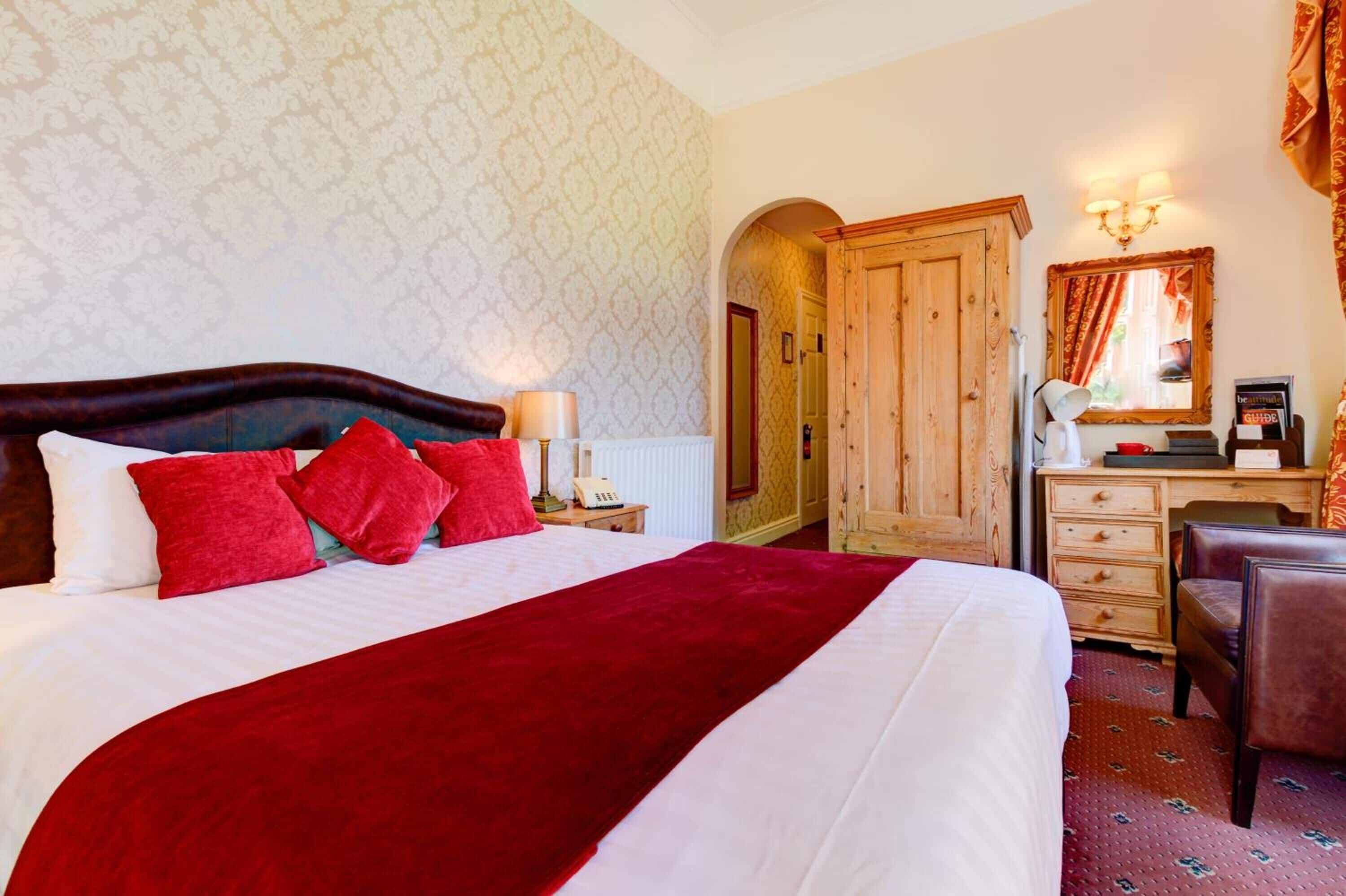 A hotel room with a large bed, red pillows, a wooden wardrobe, a desk with a lamp, and a mirror.