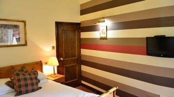 Ein Hotelzimmer mit einem Bett, einem Nachttisch mit Lampe, einem an der Wand befestigten Fernseher und einer Tür.