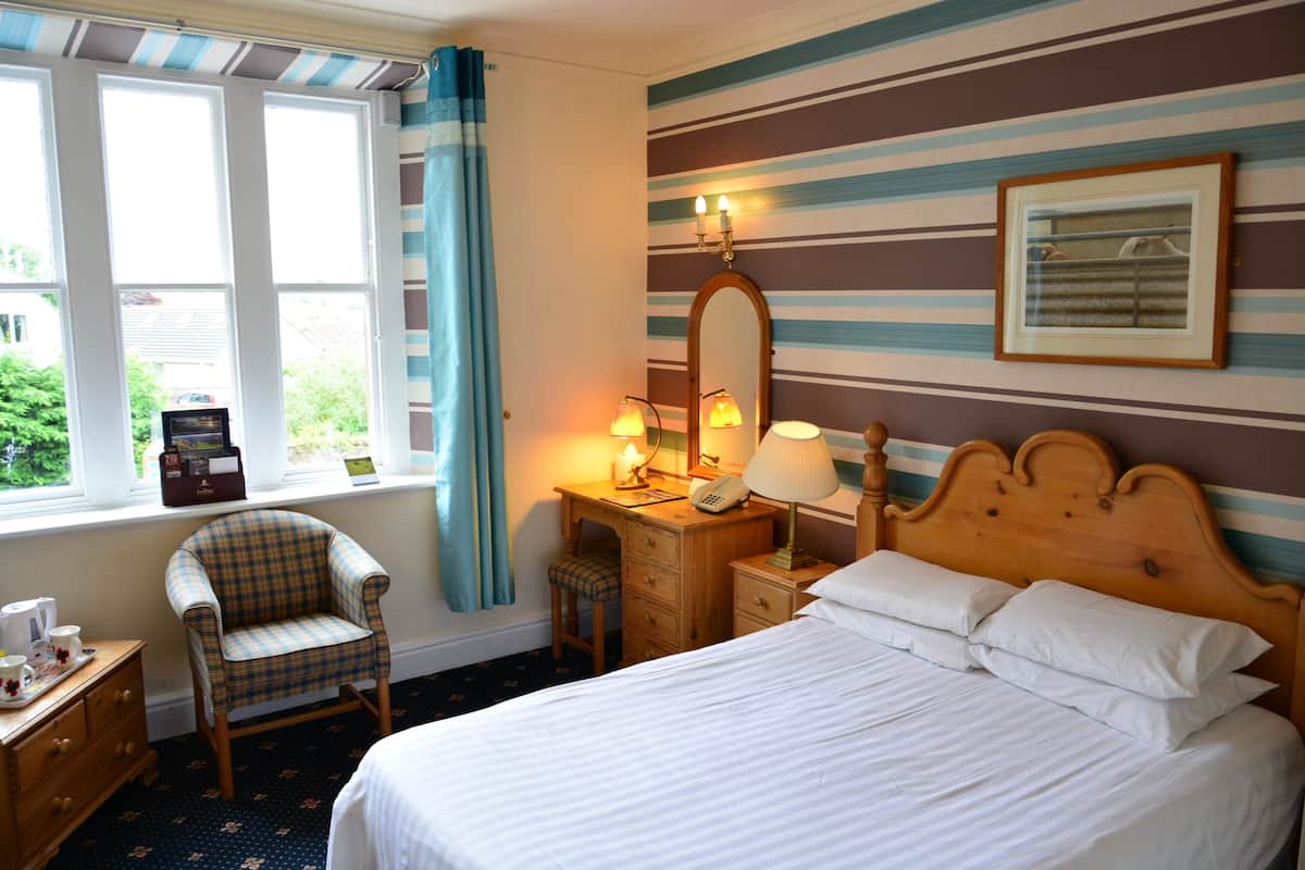 A hotel room with a bed, a wooden nightstand with a lamp, a chair, a desk with a telephone, and a window with curtains.