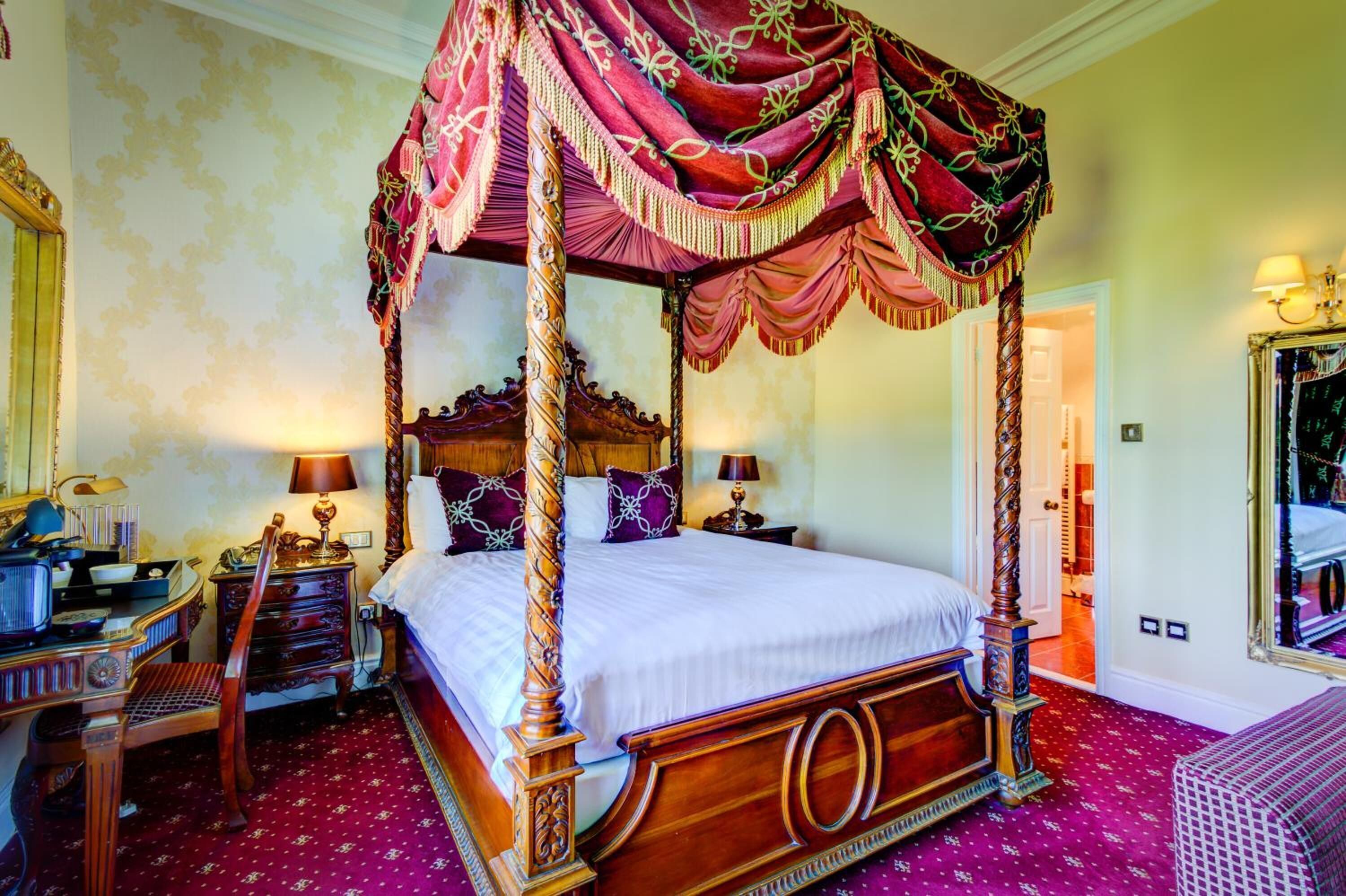 A four-poster canopy bed with ornate wooden carvings, a matching nightstand, and a red patterned carpet.