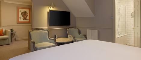 Une chambre d’hôtel avec un grand lit, deux fauteuils, une petite table, un téléviseur à écran plat fixé au mur et une porte donnant sur une salle de bain avec une douche carrelée de carreaux blancs.