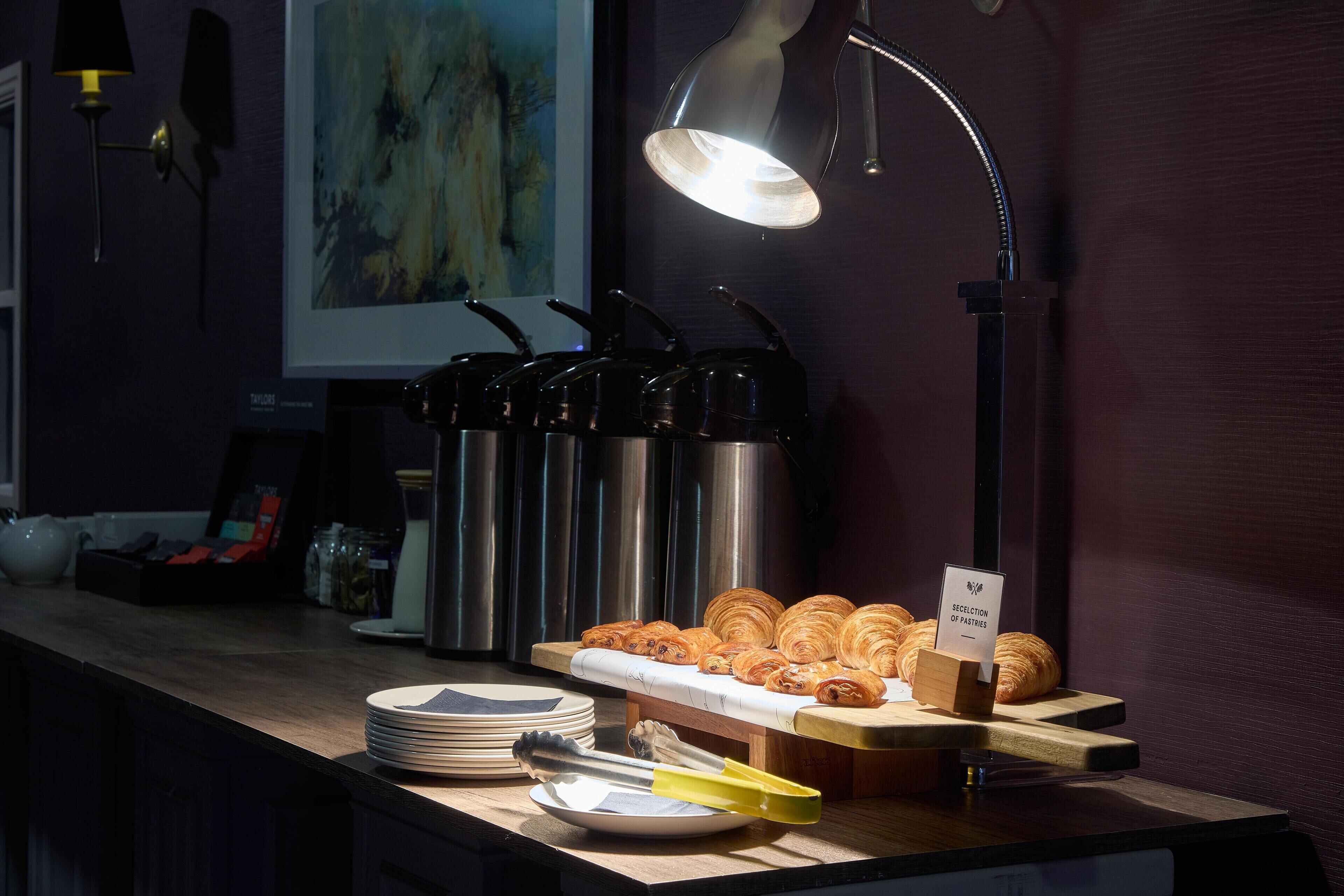 A breakfast buffet with croissants and hot beverages, a coffee machine, and a sign indicating selection of pastries.