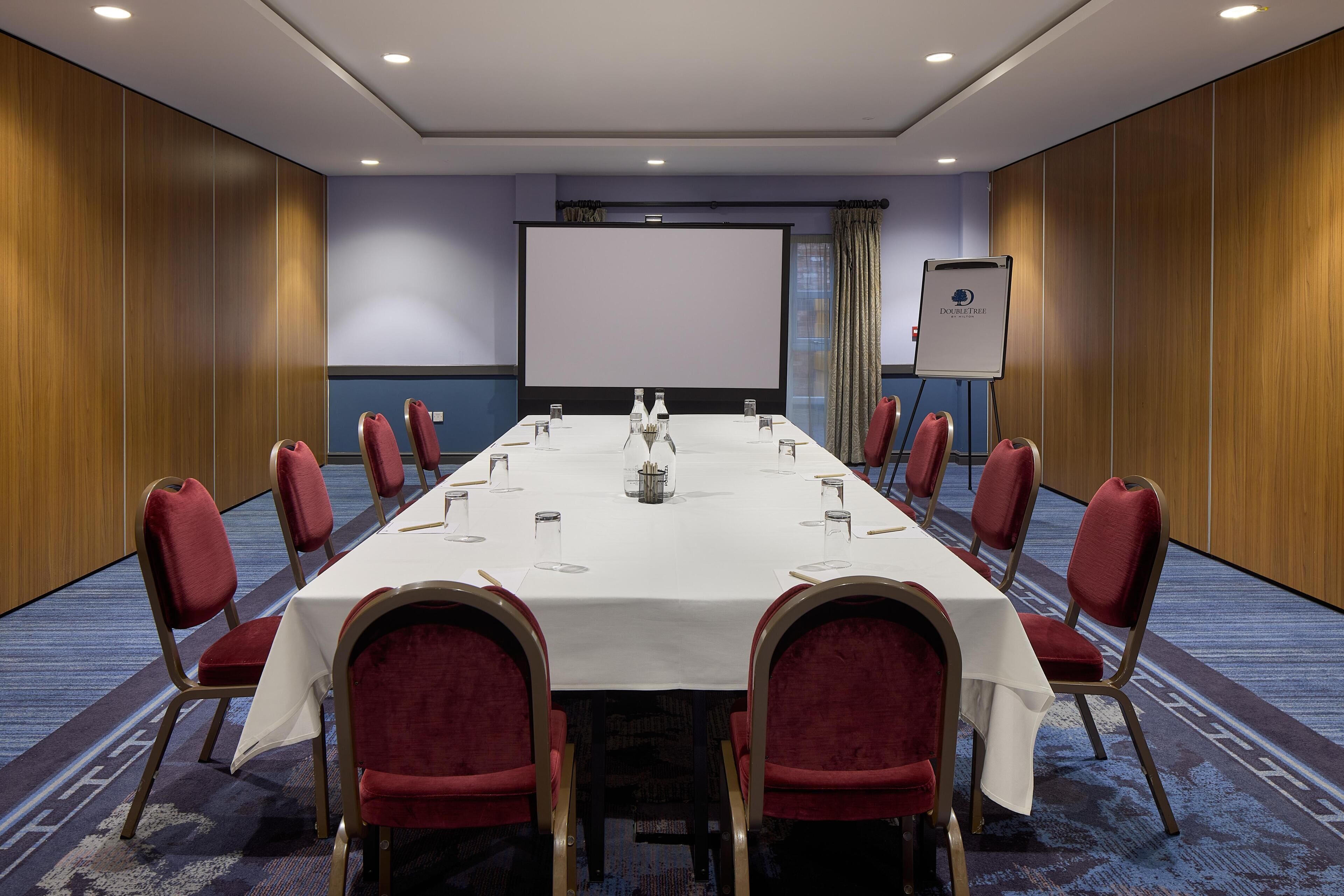 Un salón de conferencias con una larga mesa, sillas tapizadas en rojo, una pantalla de proyección blanca y un cartel de "DoubleTree".