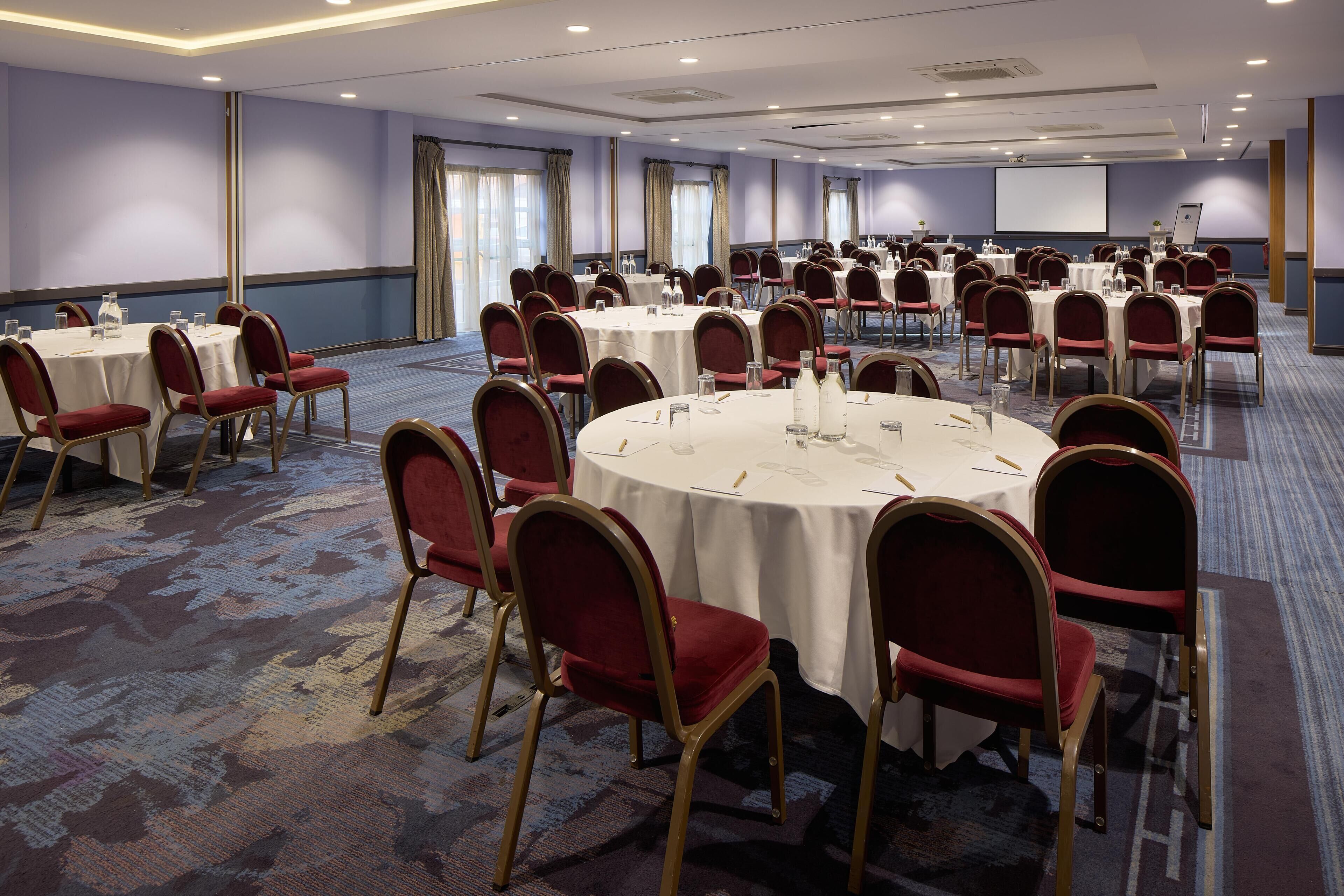 A conference room with multiple round tables, red upholstered chairs, and a large screen at the front.