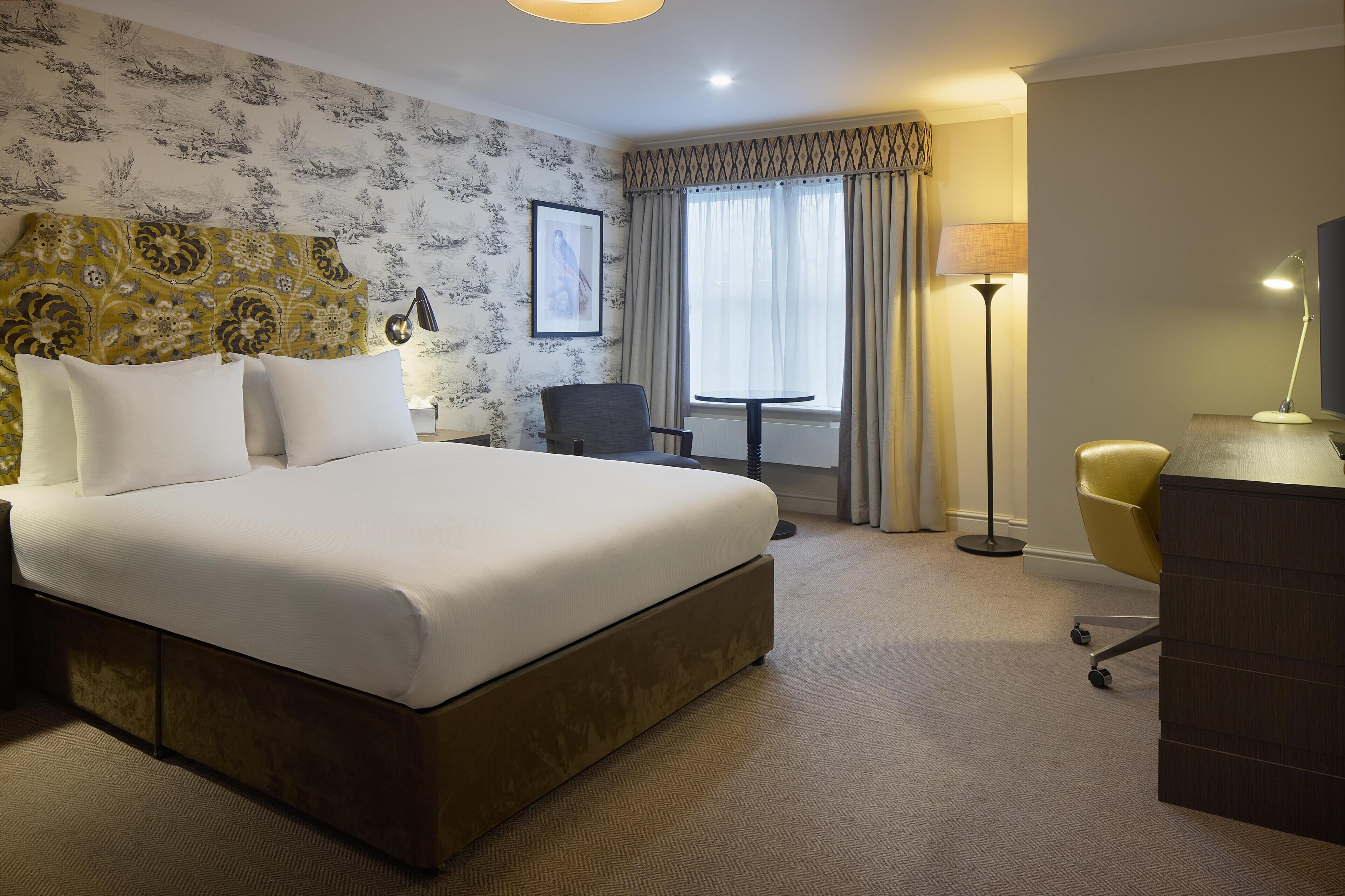 A hotel room with a large bed, a desk, a chair, a lamp, and a window with curtains.