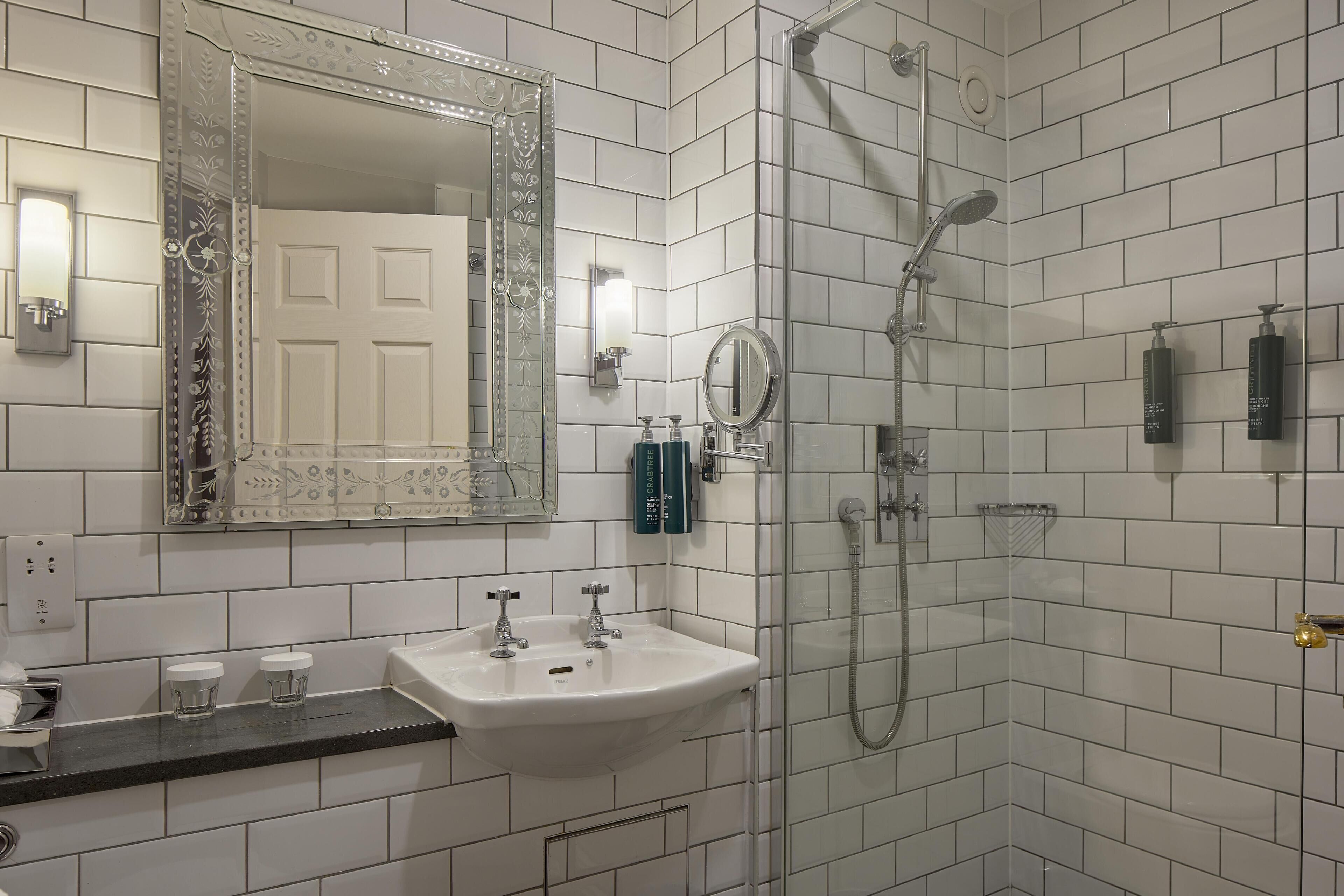 A bathroom with white tiled walls, a sink with two faucets, a large mirror, a shower with a handheld showerhead, and two bottles of shampoo on the wall.