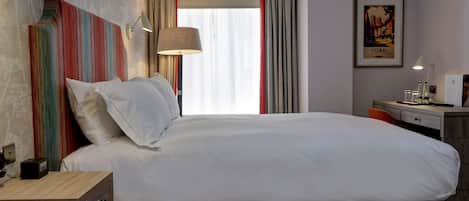 Quarto de hotel com uma cama grande, uma mesinha de cabeceira com abajur, uma escrivaninha com cadeira e um quadro pendurado na parede.