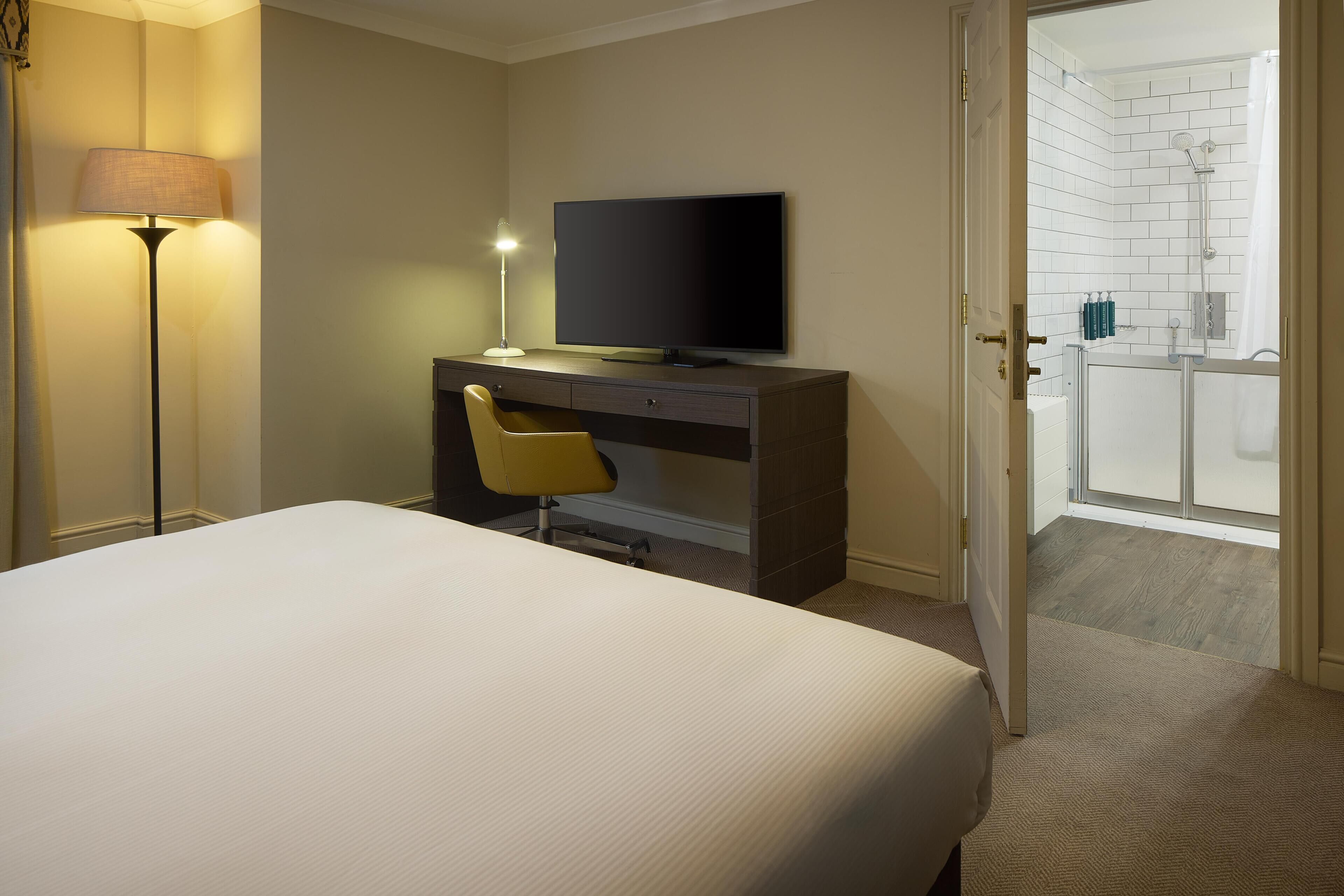 Habitación de hotel con una cama, un escritorio con televisor, una silla, una lámpara y una puerta que da a un baño con ducha.