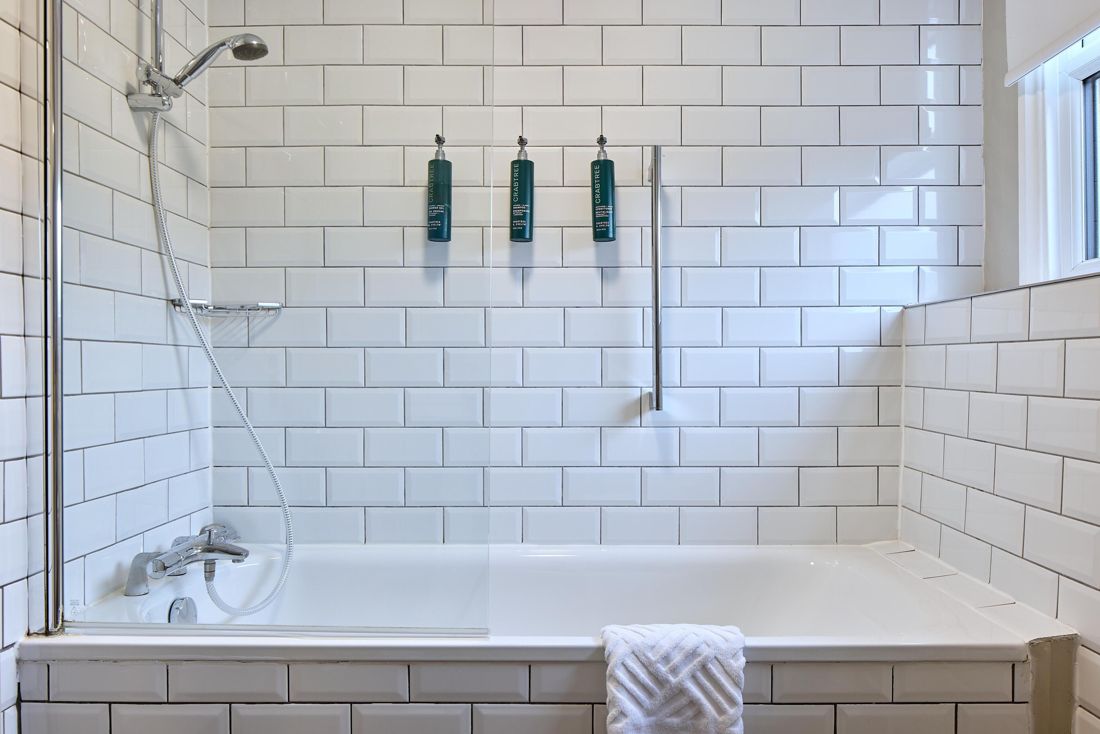 A bathroom with a white tiled wall, a bathtub, a showerhead, and three bottles of Crave body wash.
