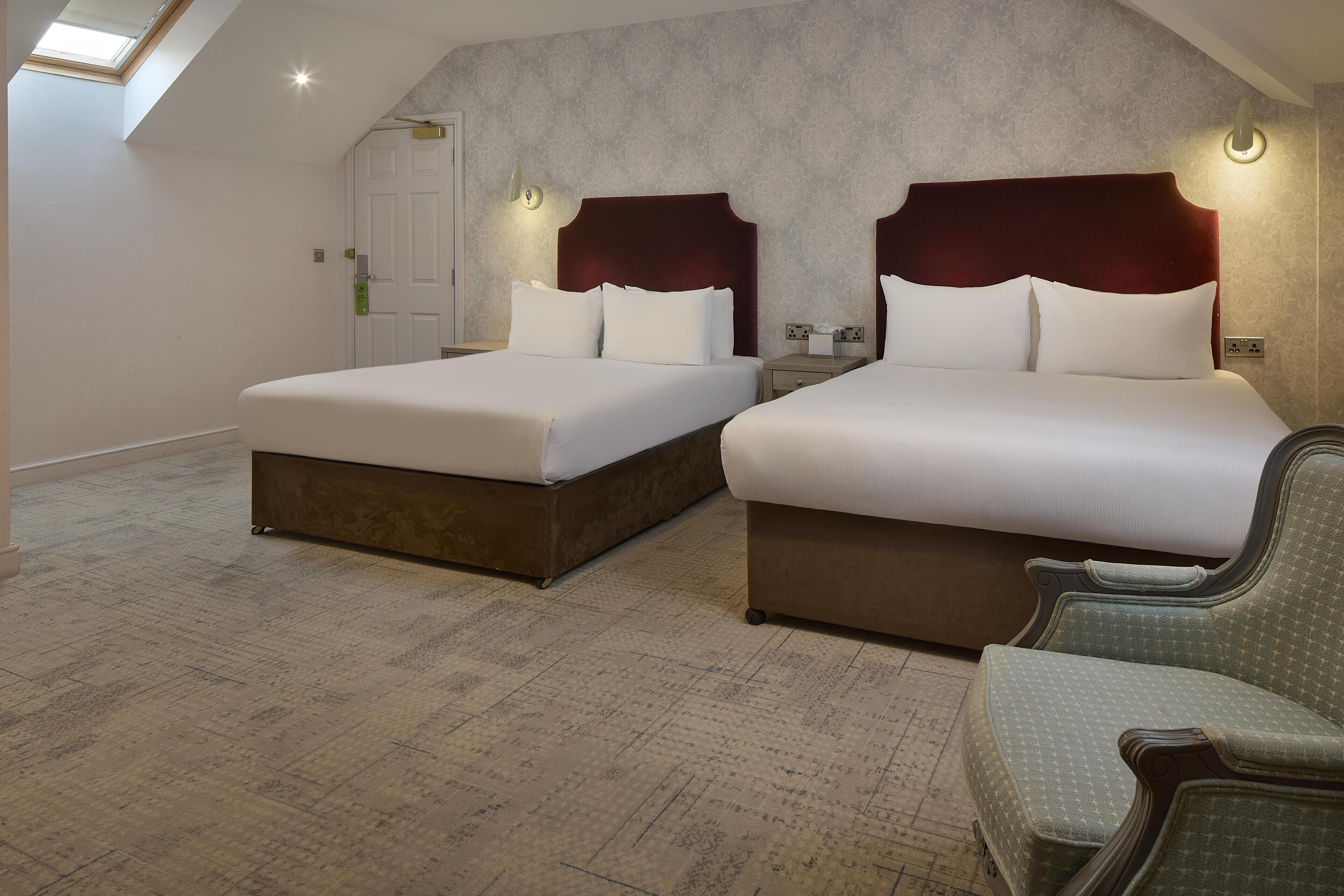A hotel room with two beds, a skylight, a patterned carpet, and a green upholstered chair.