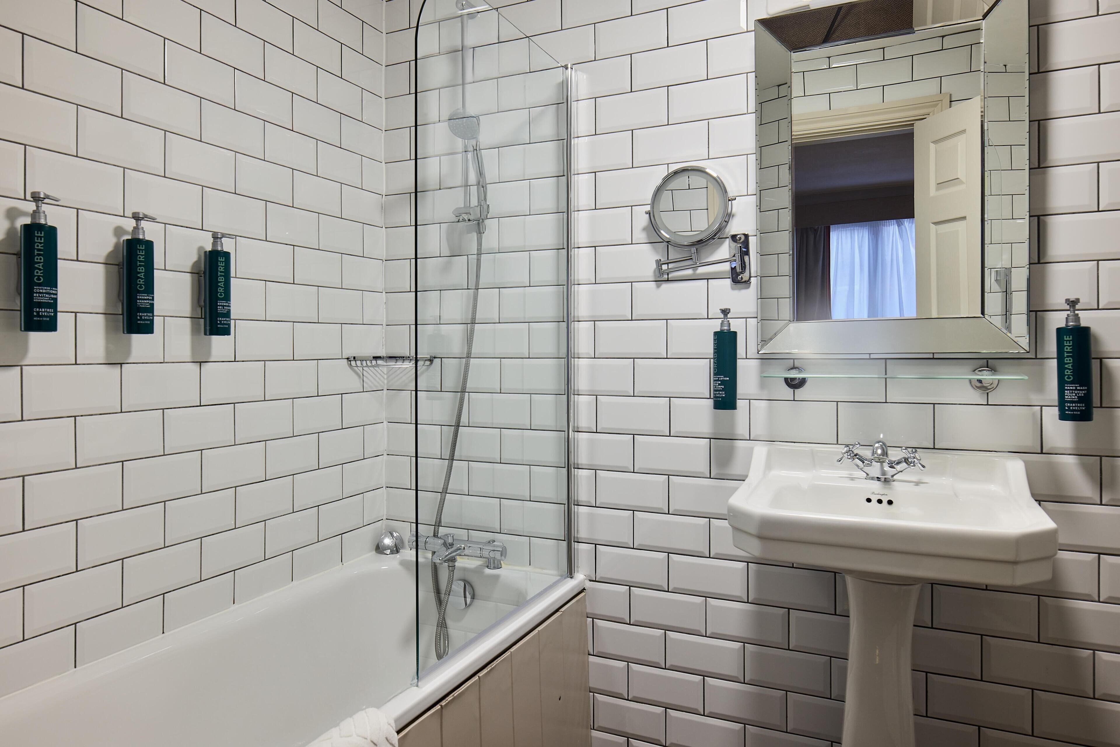 A bathroom with white tiled walls, a glass-enclosed shower, a pedestal sink, and three bottles of Craytree body wash on the wall.