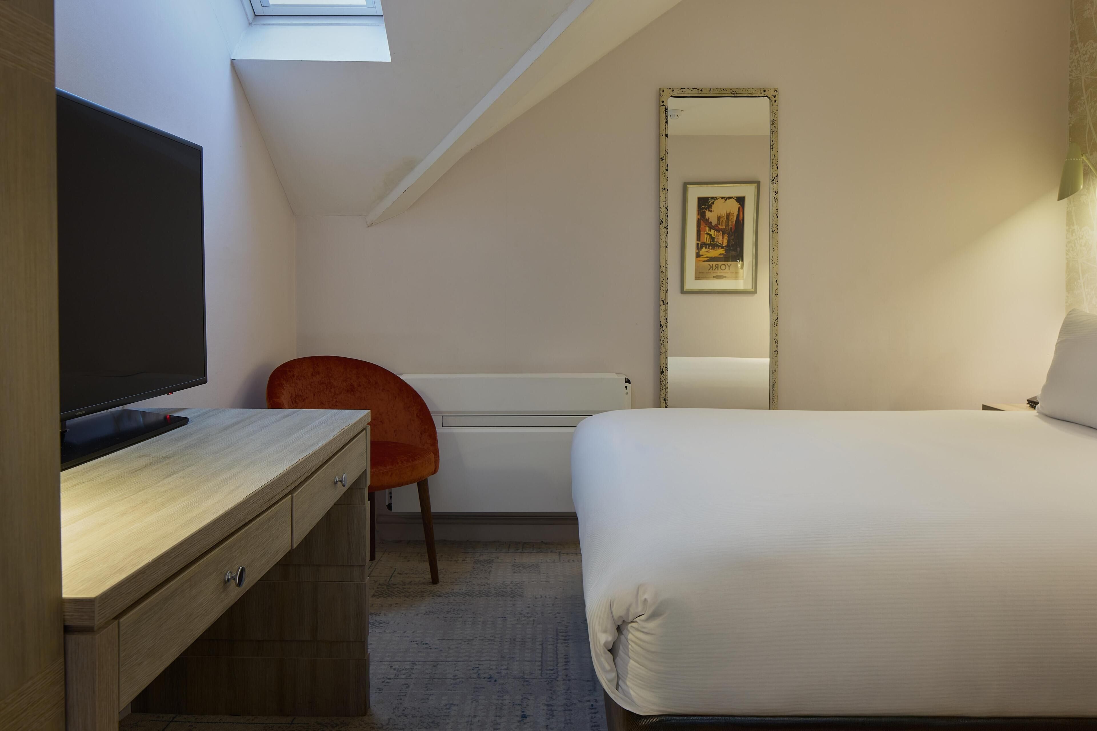 Un dormitorio con cama, escritorio con televisor, una silla, radiador y un cuadro en la pared.