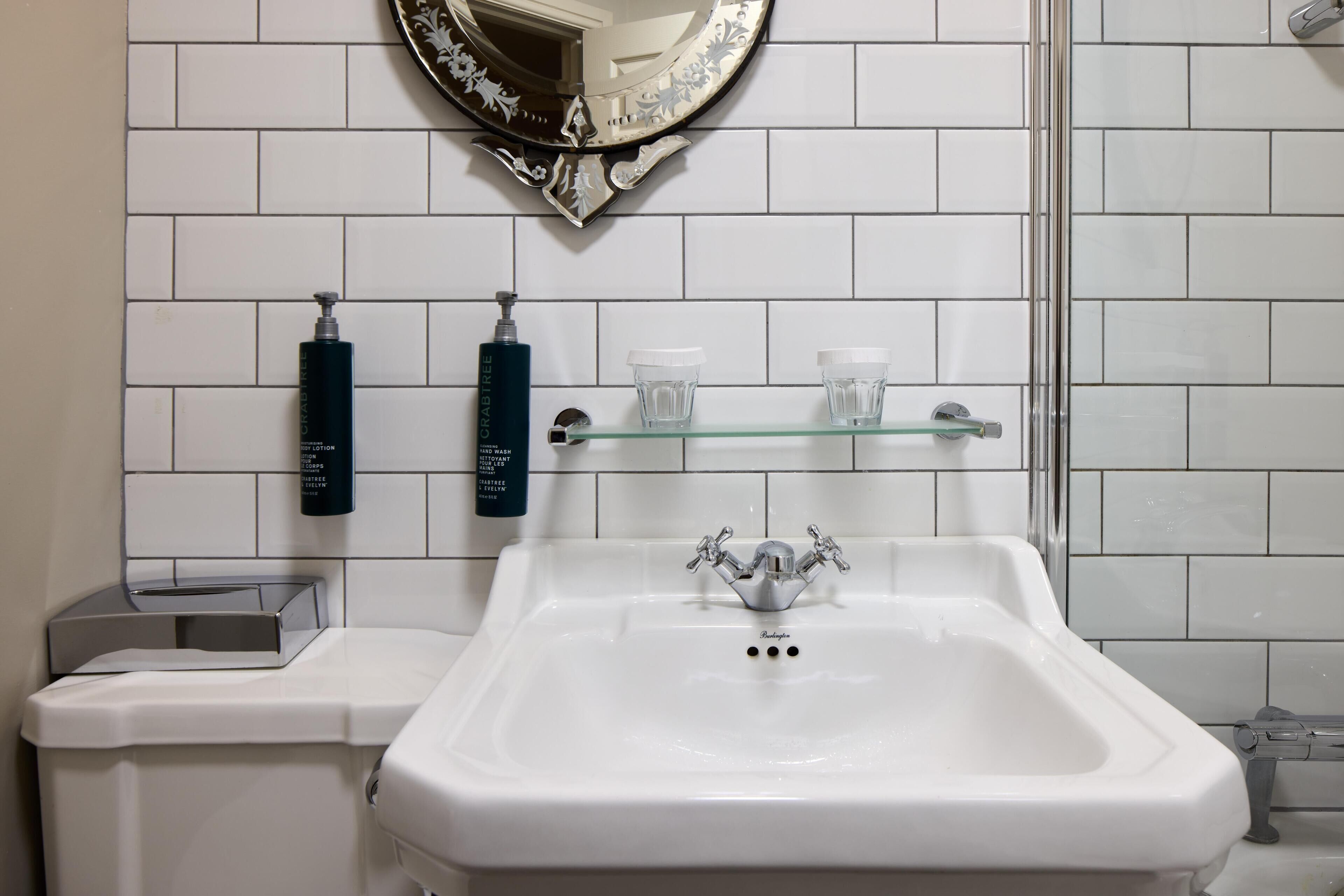 A bathroom with a white sink, two bottles of hand soap, a glass shelf with cups, and a toilet.