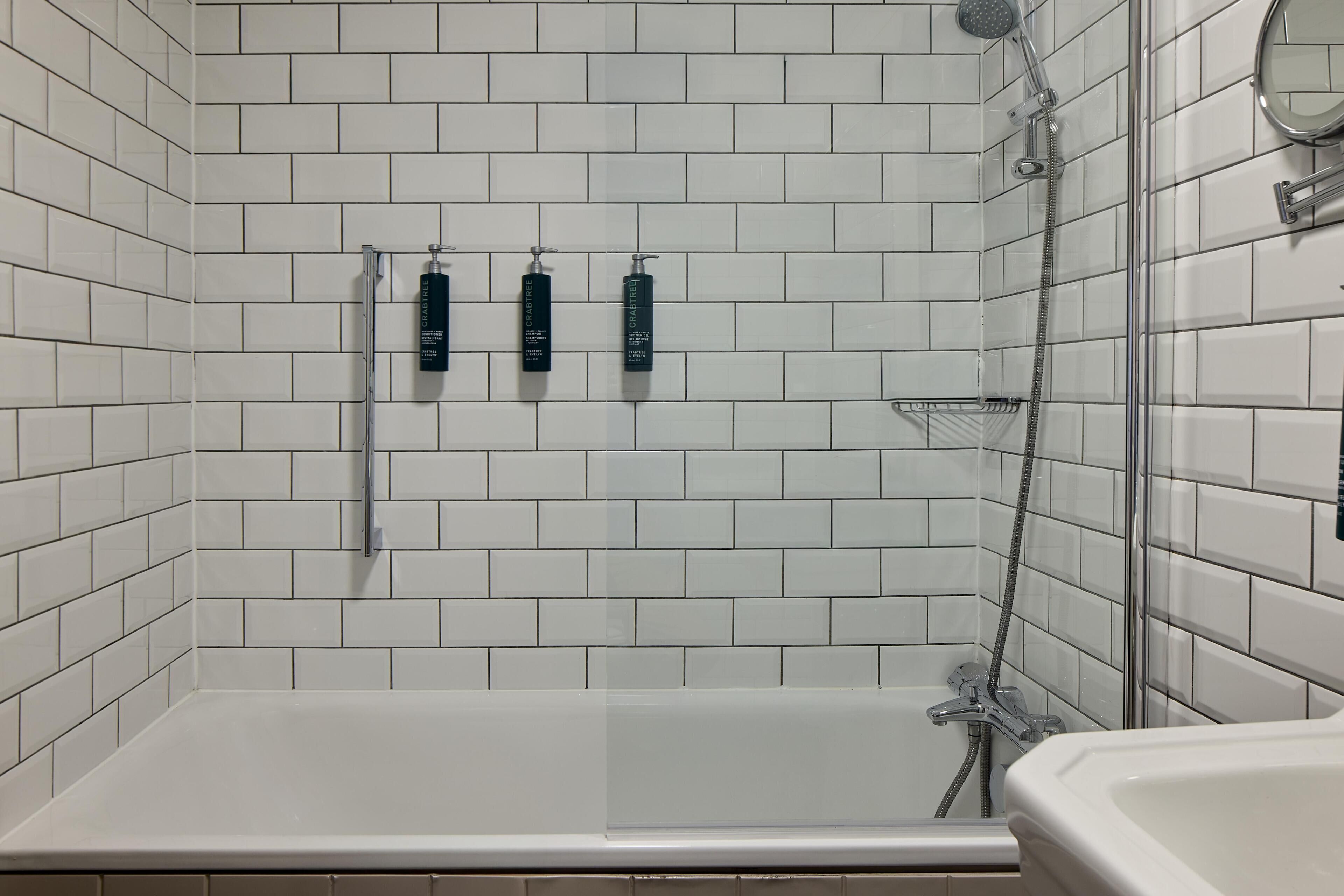 Un baño con paredes de azulejos blancos, una bañera y tres botellas de champú en la pared.