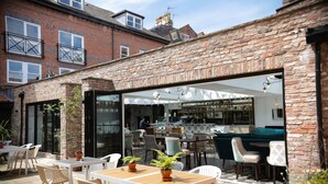 Un patio exterior con mesas y sillas, un muro de ladrillo y una puerta de cristal que da a un espacio interior con asientos y una zona de bar.