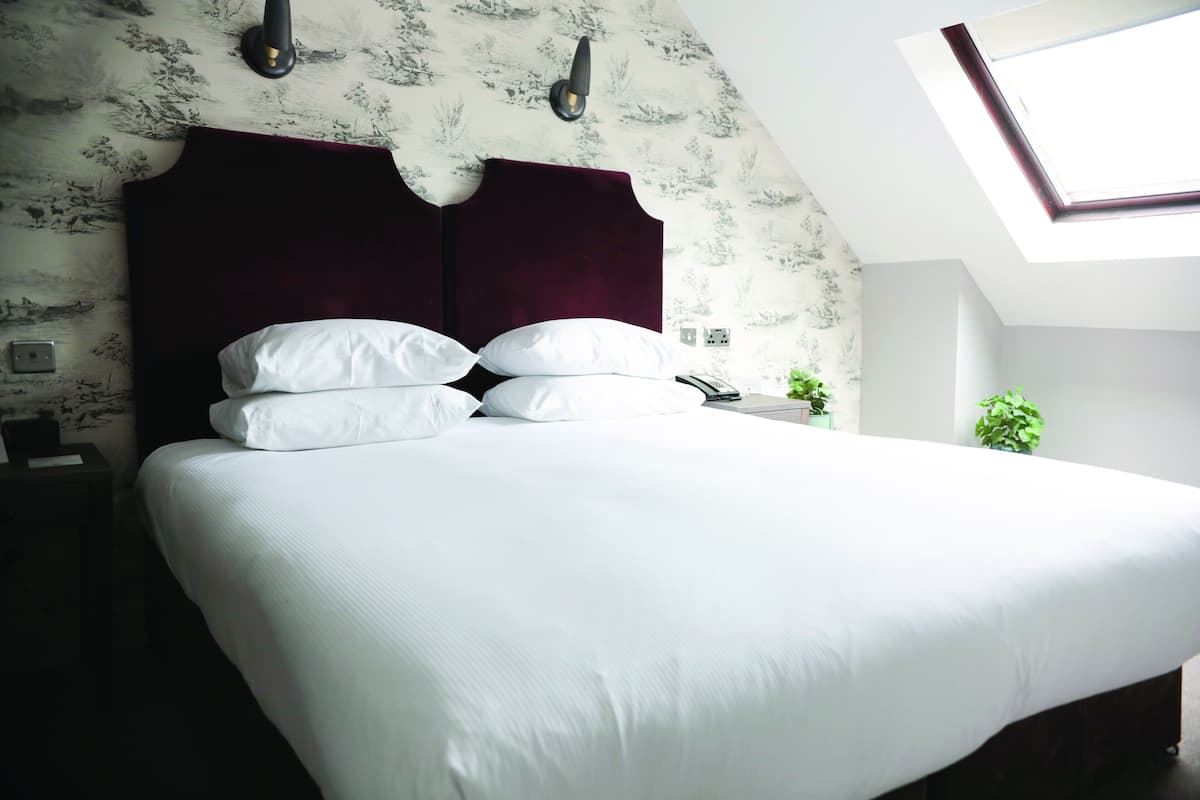 En snyggt bäddad säng med vita sängkläder och en mörk sänggavel i ett hotellrum med ett takfönster och tapet med mönster.