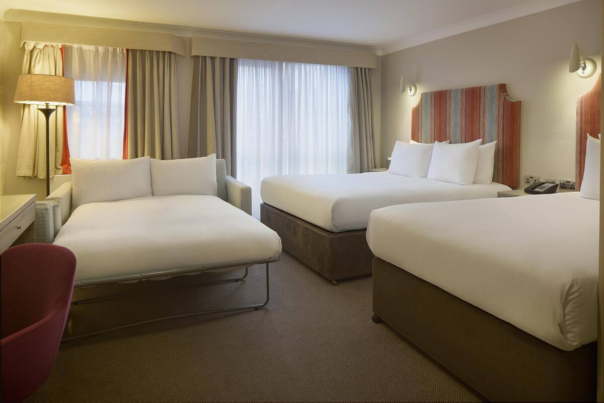 Ett hotellrum med två sängar, ett skrivbord, en stol, en lampa och ett fönster med gardiner.