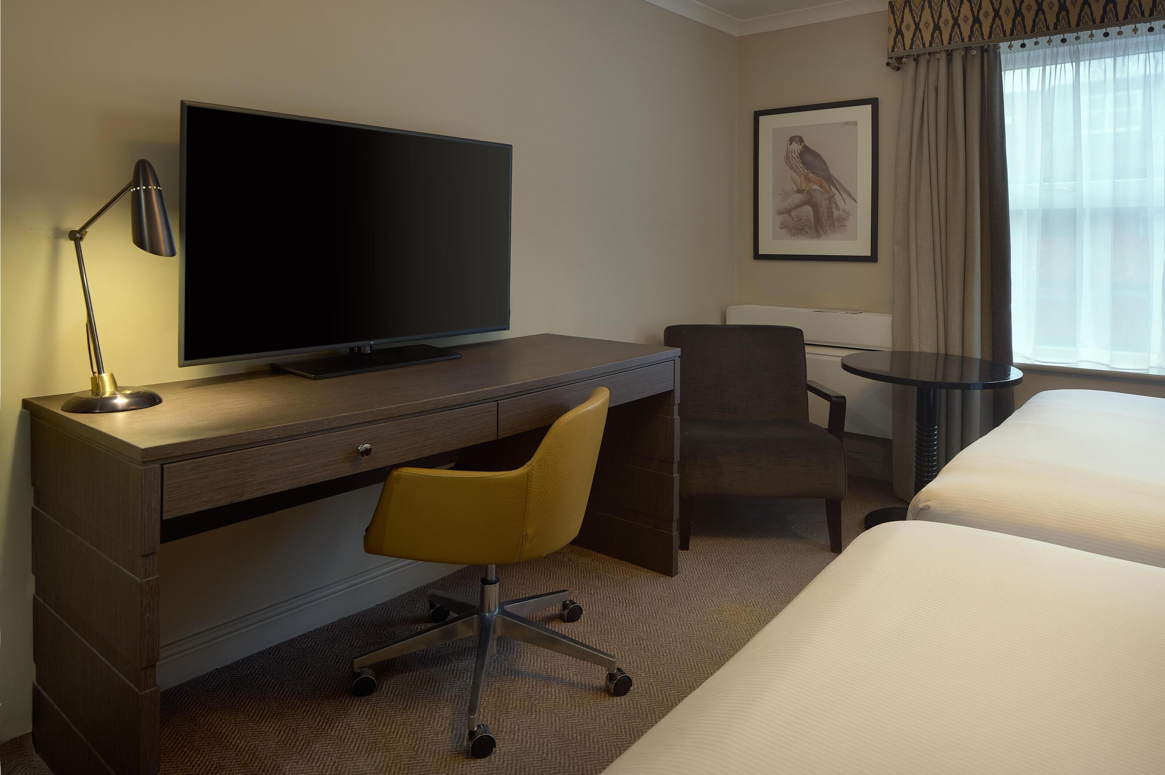 Quarto de hotel com televisão de tela plana sobre uma mesa de madeira, uma cadeira de escritório amarela, uma pequena mesa redonda, uma cama e um quadro pendurado na parede.