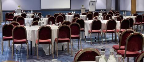 Un salón de conferencias con varias mesas, sillas rojas y una pantalla de proyector.