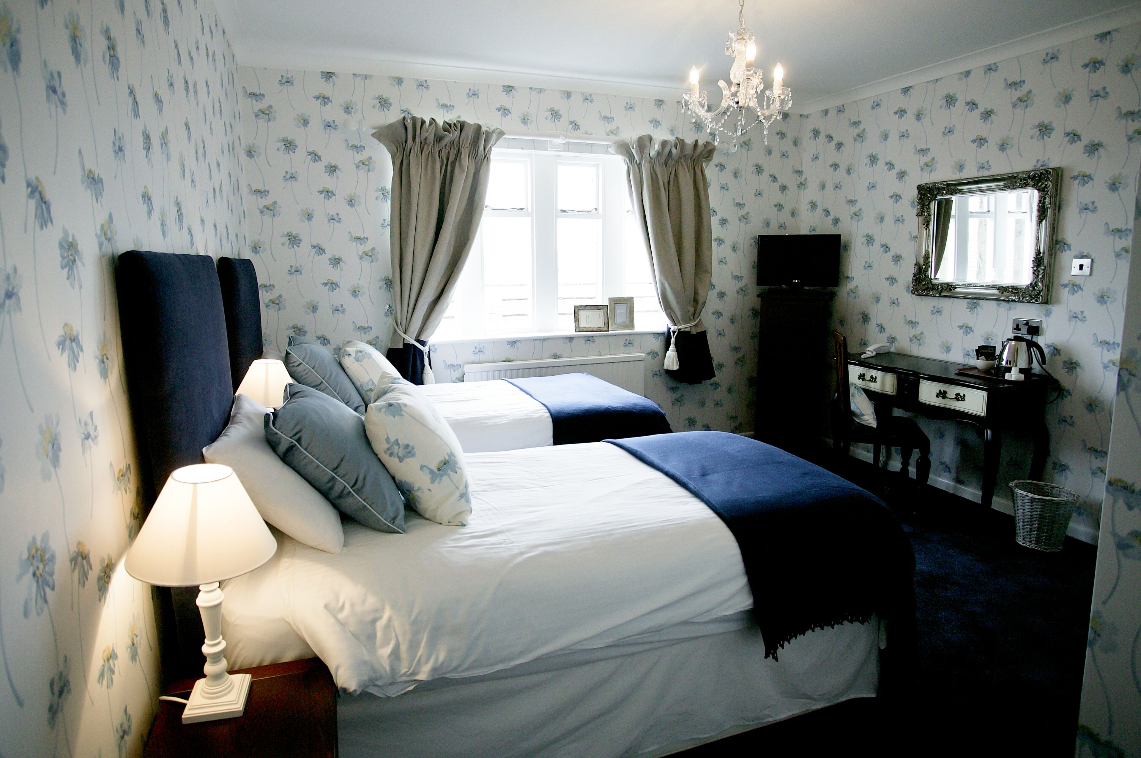 Une chambre avec deux lits, une commode, un miroir, un lustre et une fenêtre avec des rideaux.
