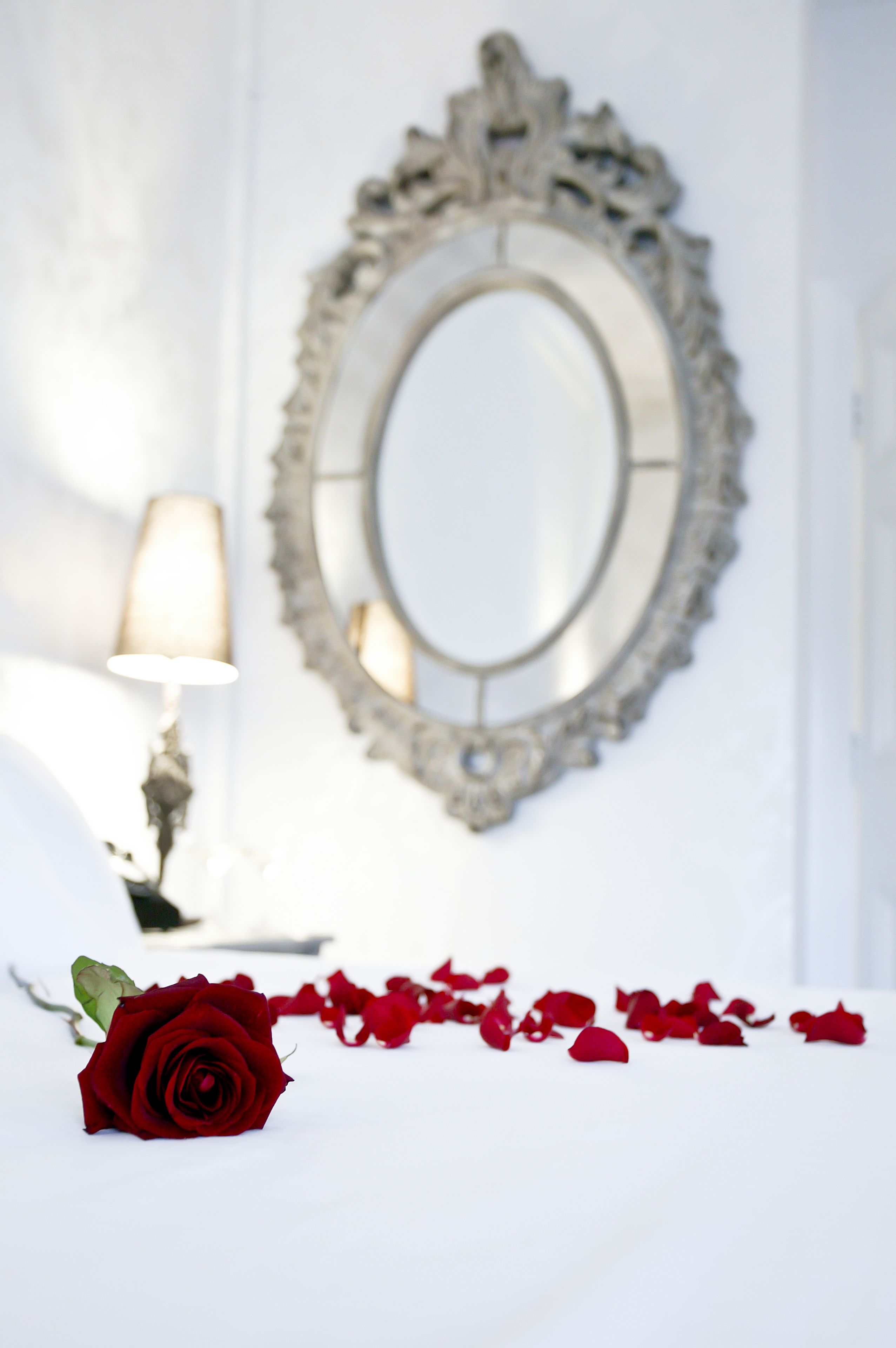 Una habitación blanca con un espejo grande y ornamentado, una lámpara de noche y una sola rosa roja sobre la cama.