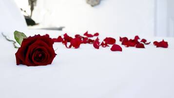 Una habitación blanca con un espejo grande y ornamentado, una lámpara de noche y una sola rosa roja sobre la cama.