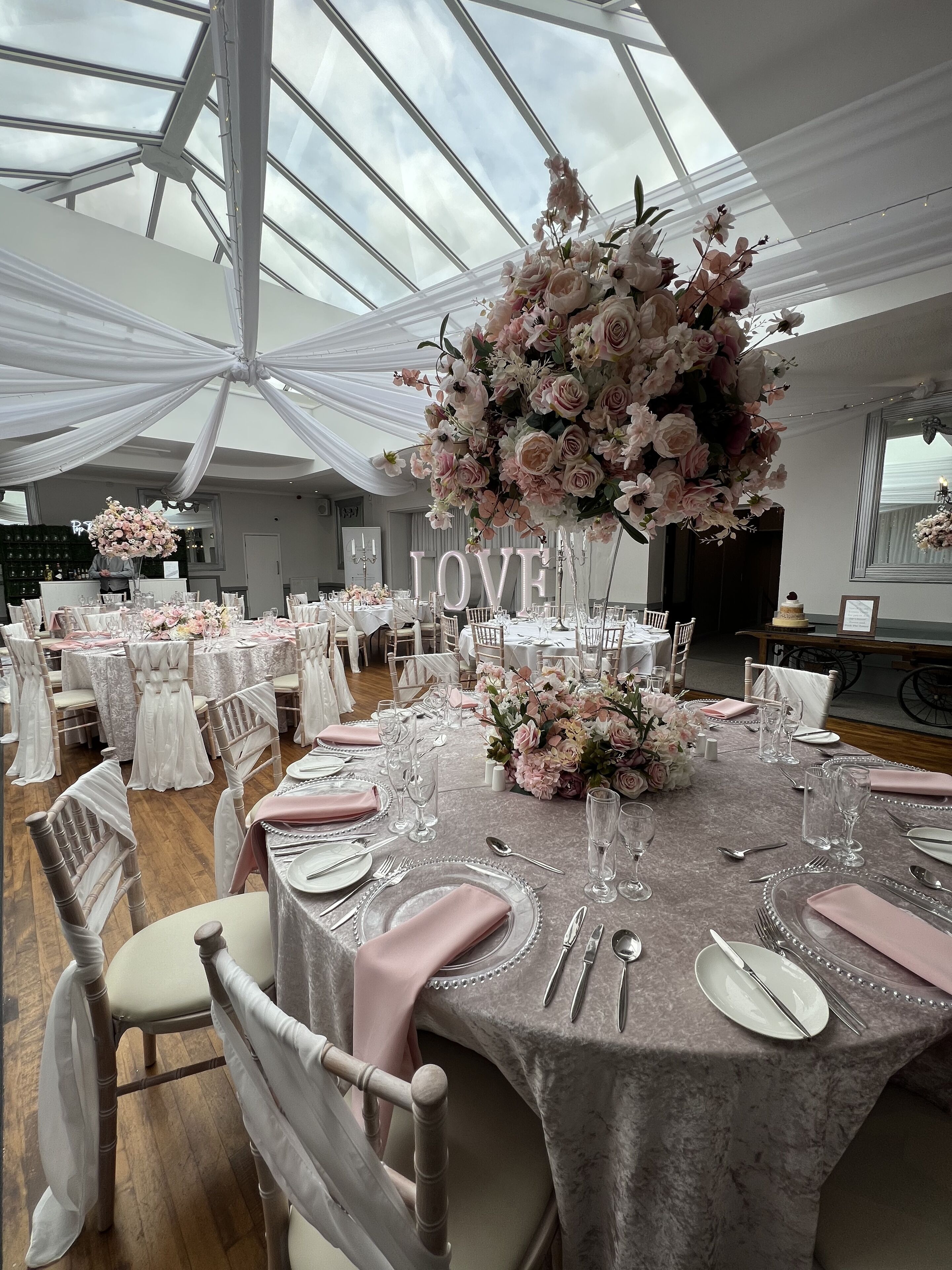 A banquet hall with round tables set for a formal event, featuring white and pink floral centerpieces and a large 'LOVE' sign.