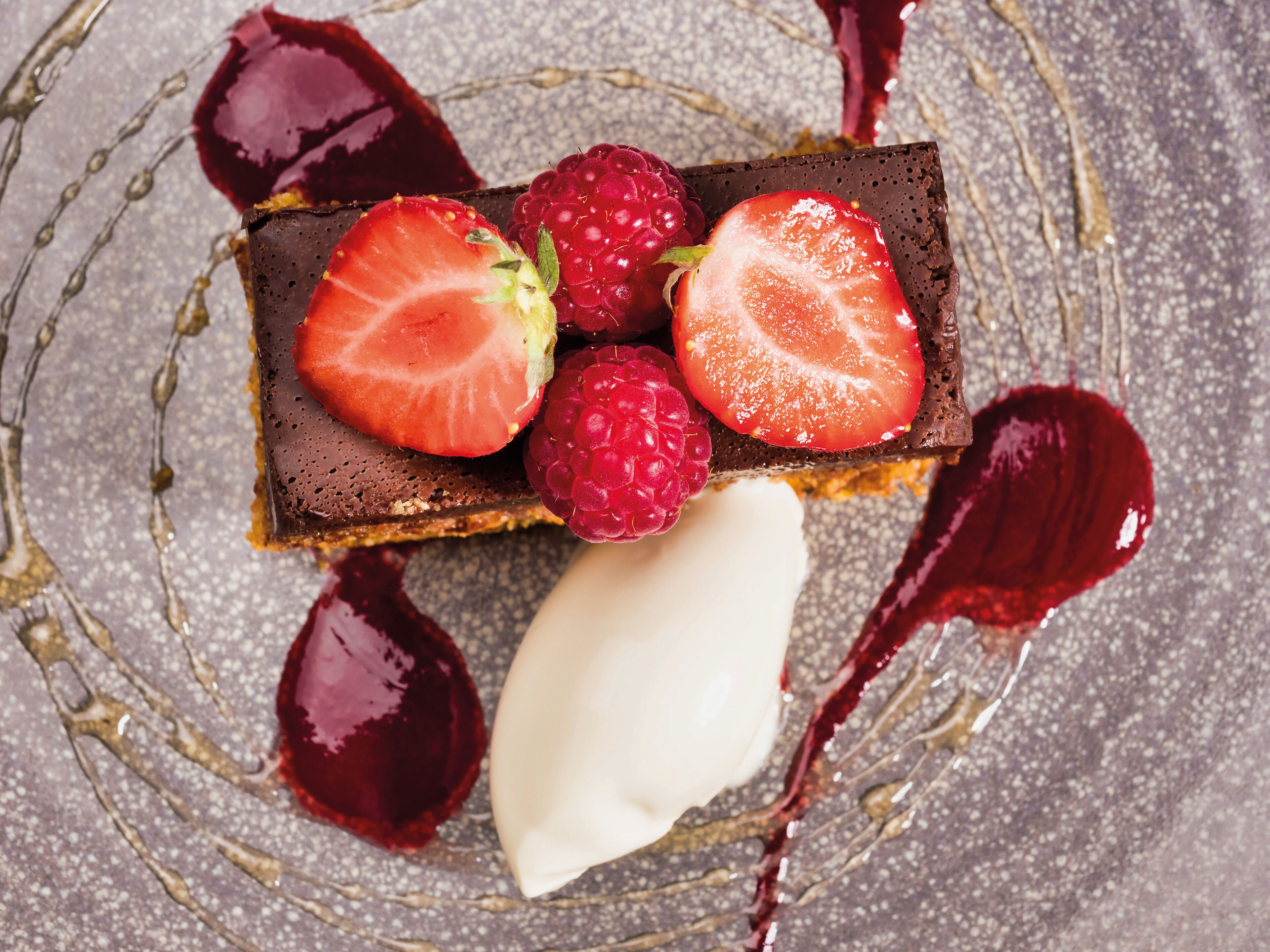 Un postre con fresas y frambuesas sobre una base de chocolate, acompañado de una bola de helado y salsa roja.