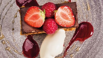 Un postre con fresas y frambuesas sobre una base de chocolate, acompañado de una bola de helado y salsa roja.
