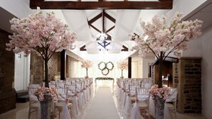 A wedding aisle with white chairs and a white carpet, flanked by large vases with pink flowers.