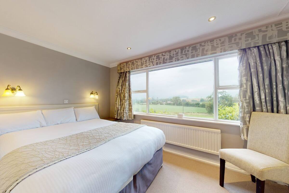 A bedroom with a large bed, two bedside lamps, a window with curtains, and a chair.