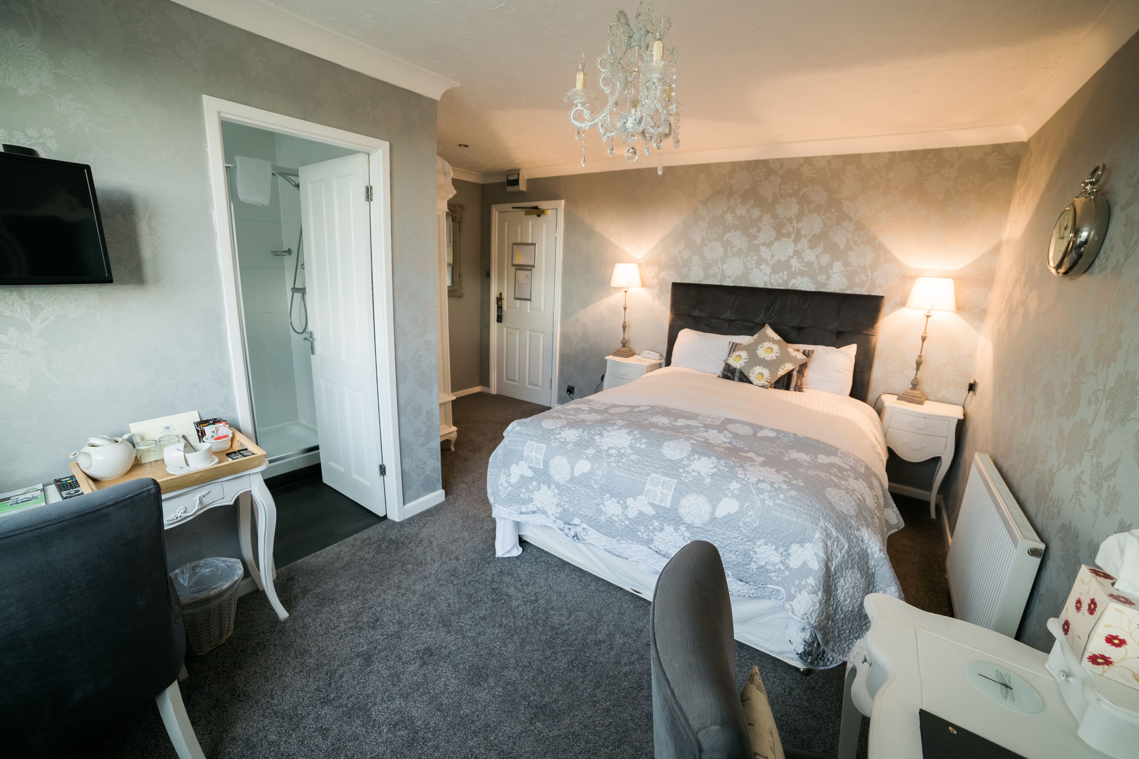 A hotel room with a bed, two bedside tables, a desk with a chair, a television, a chandelier, and a door leading to a bathroom.