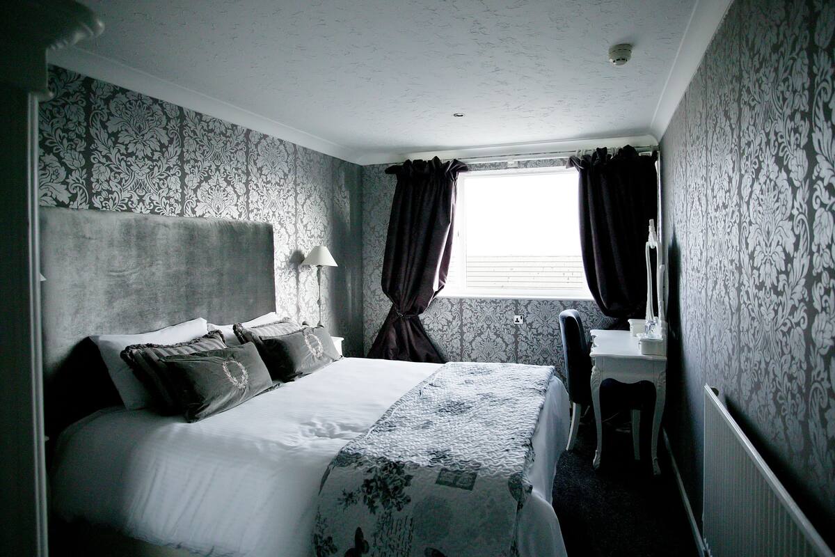 A bedroom with a bed, a bedside table, a chair, a window with curtains, and patterned wallpaper.