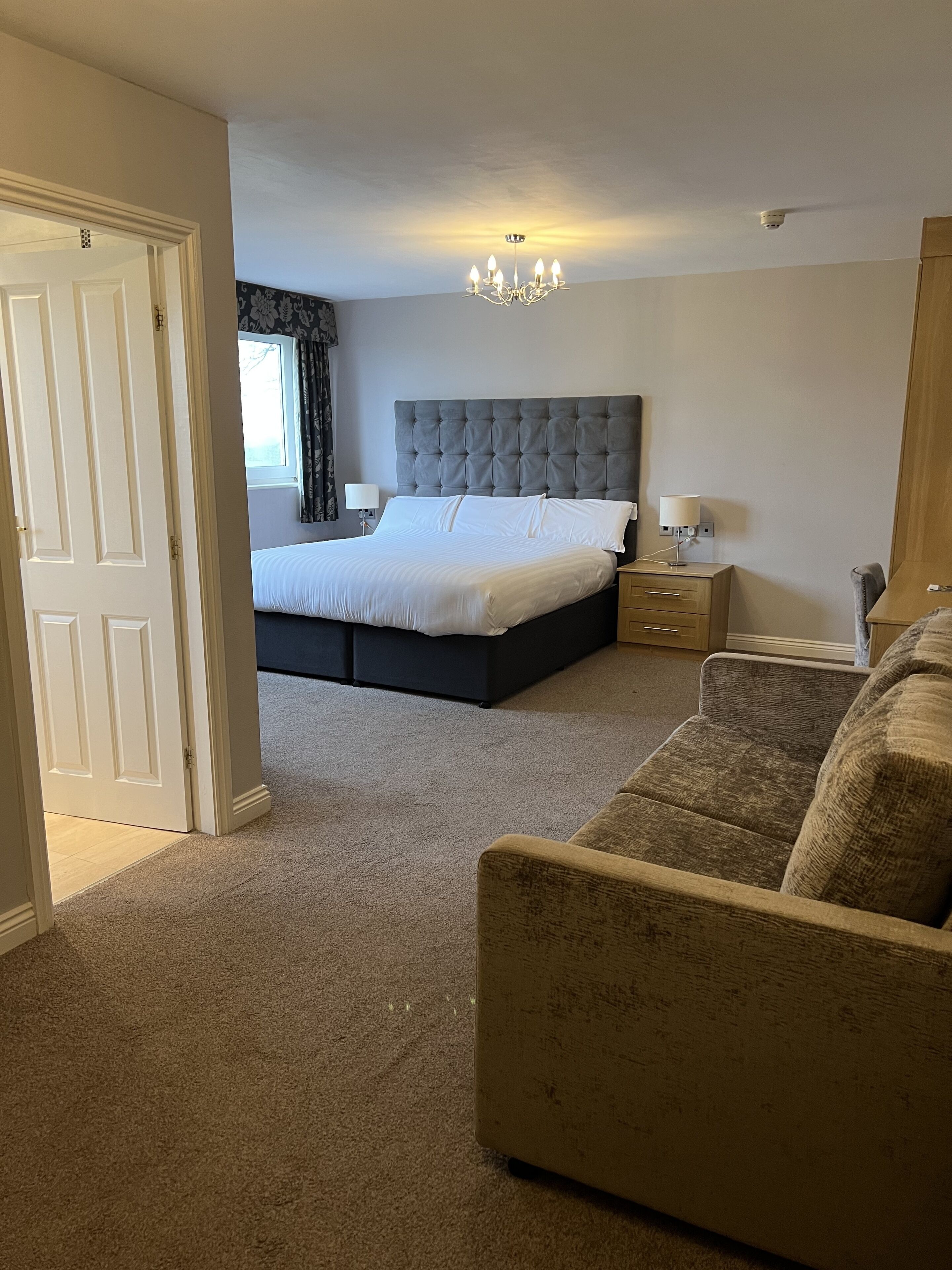 A bedroom with a large bed, a nightstand, a lamp, a chair, and a door leading to another room.