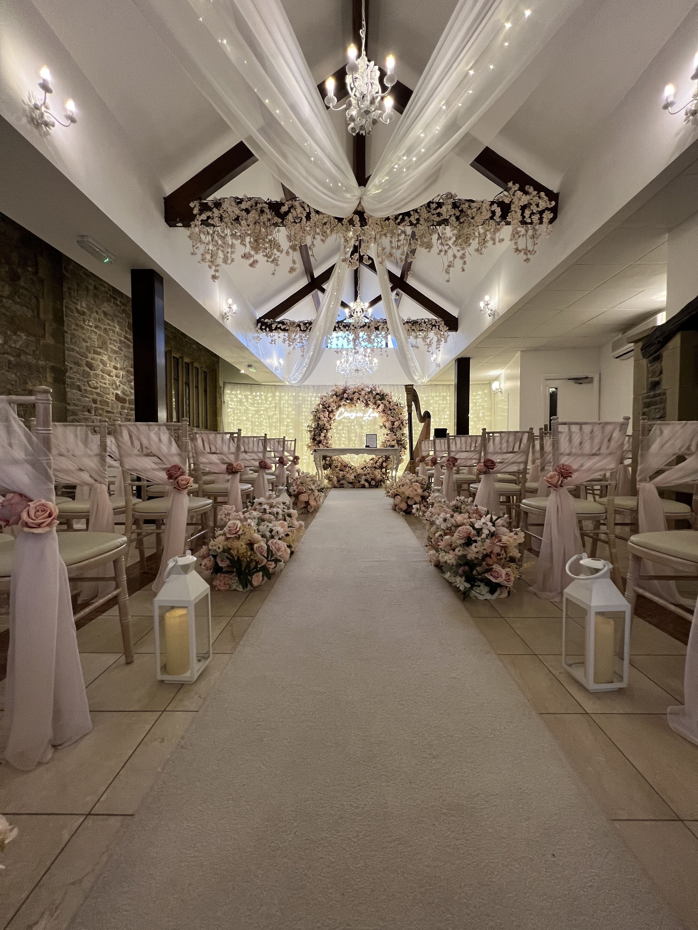 Une allée décorée pour un mariage, agrémentée de compositions florales, de chaises blanches et d’un lustre.