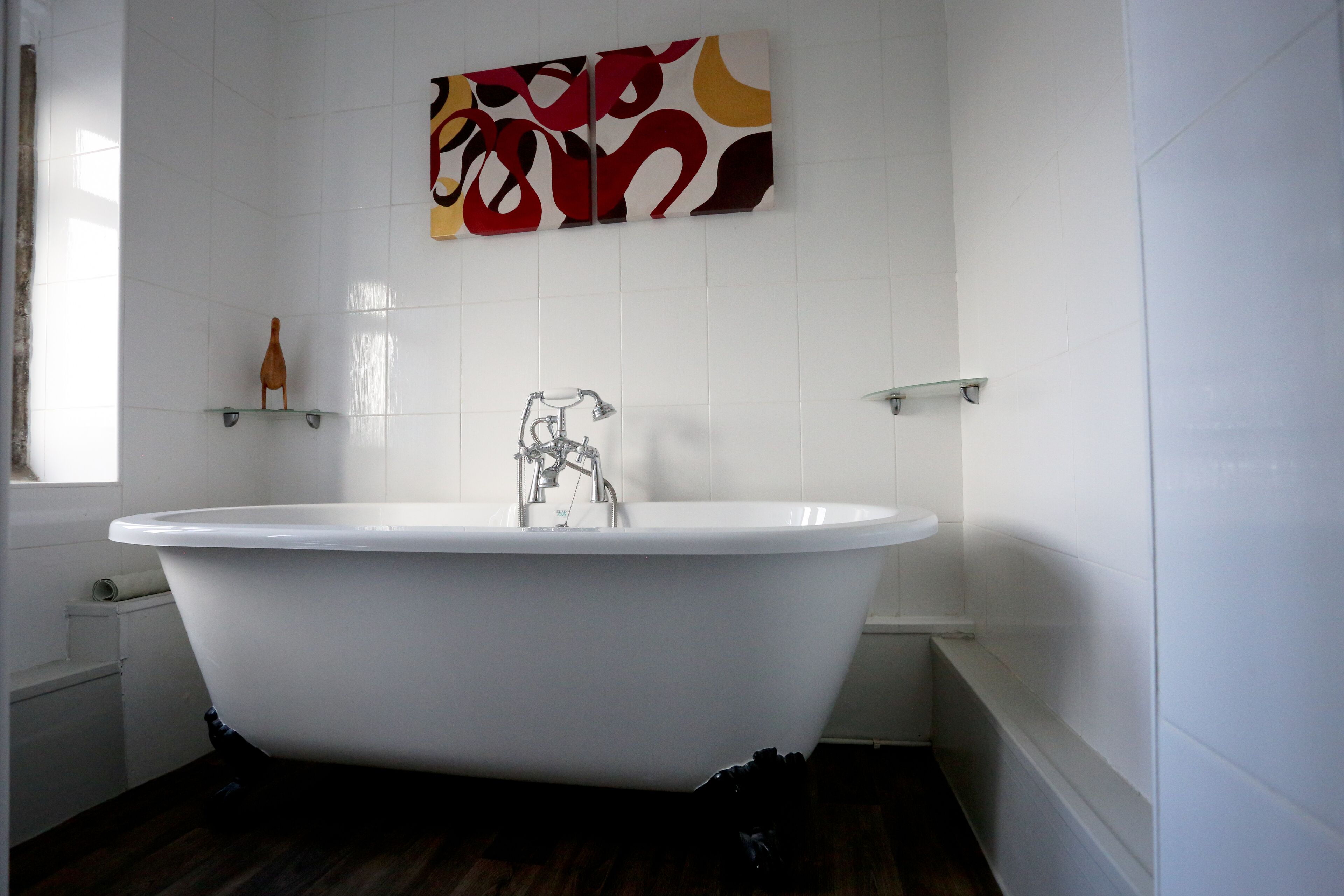A bathroom with a freestanding bathtub, white tiled walls, and a colorful abstract painting above the tub.