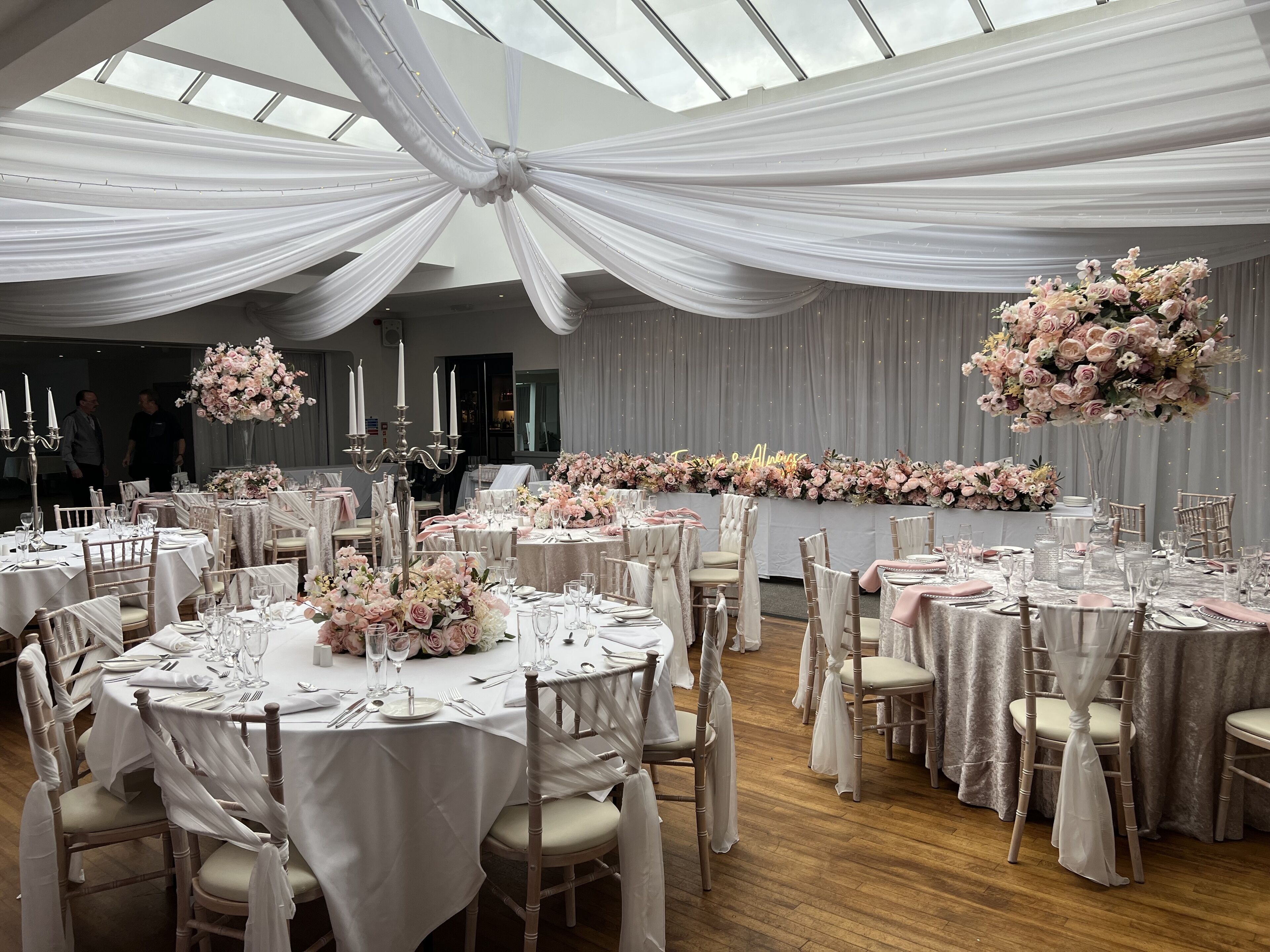 Uma sala de banquetes com mesas redondas preparadas para refeições, tetos cobertos por tecido branco e arranjos florais.