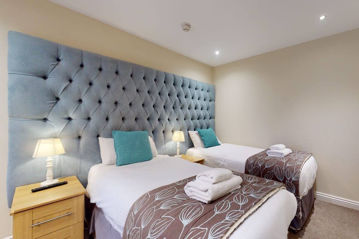 A hotel room with a tufted headboard, two beds, bedside lamps, and a radiator.