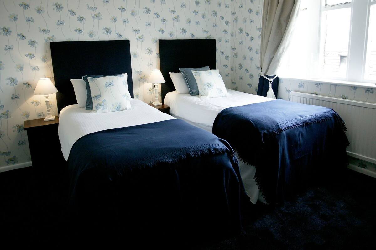 A bedroom with two beds, nightstands, a window with curtains, and floral wallpaper.