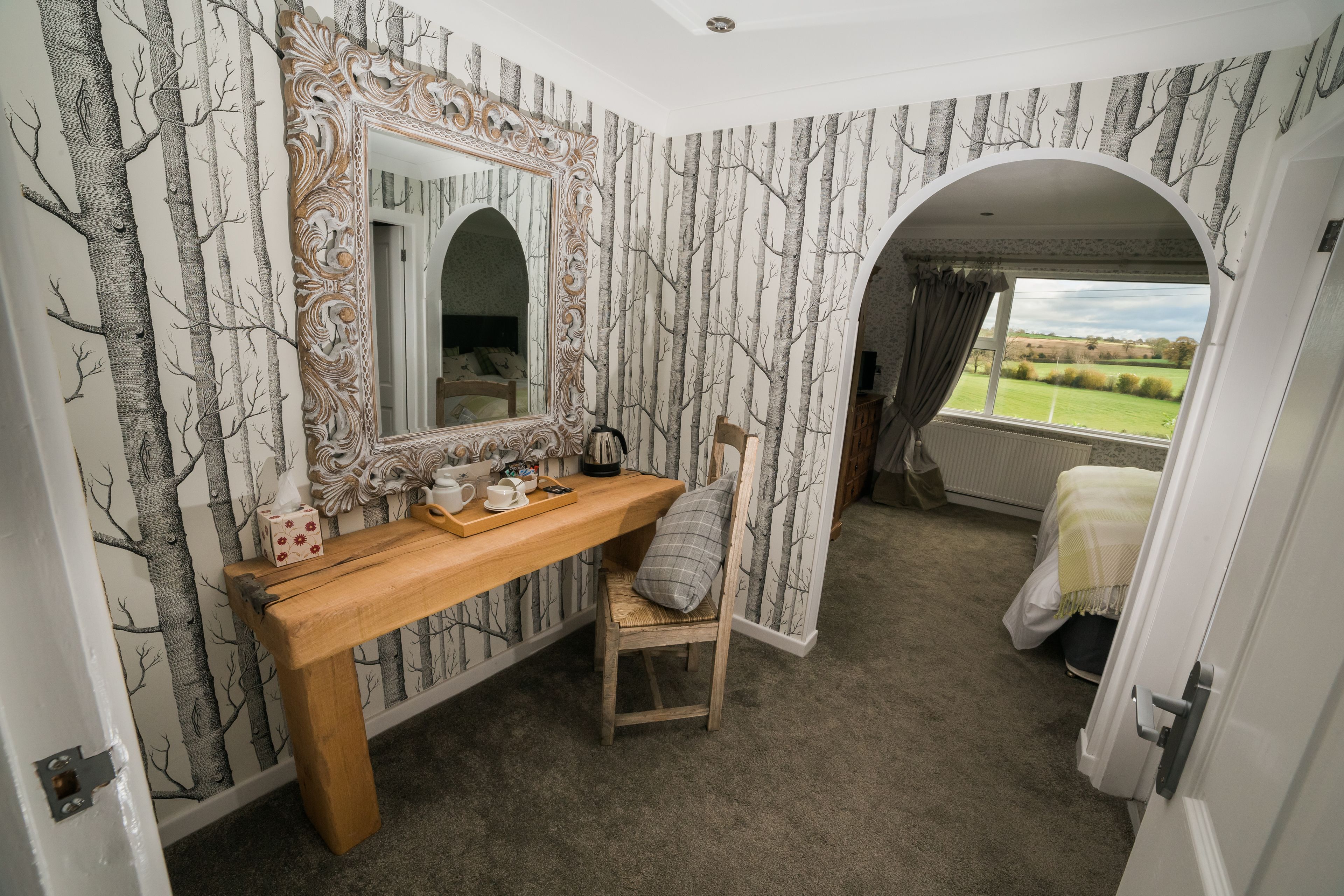A bedroom with a large mirror, a wooden bench, a chair, and a view of the outdoors through an arched window.