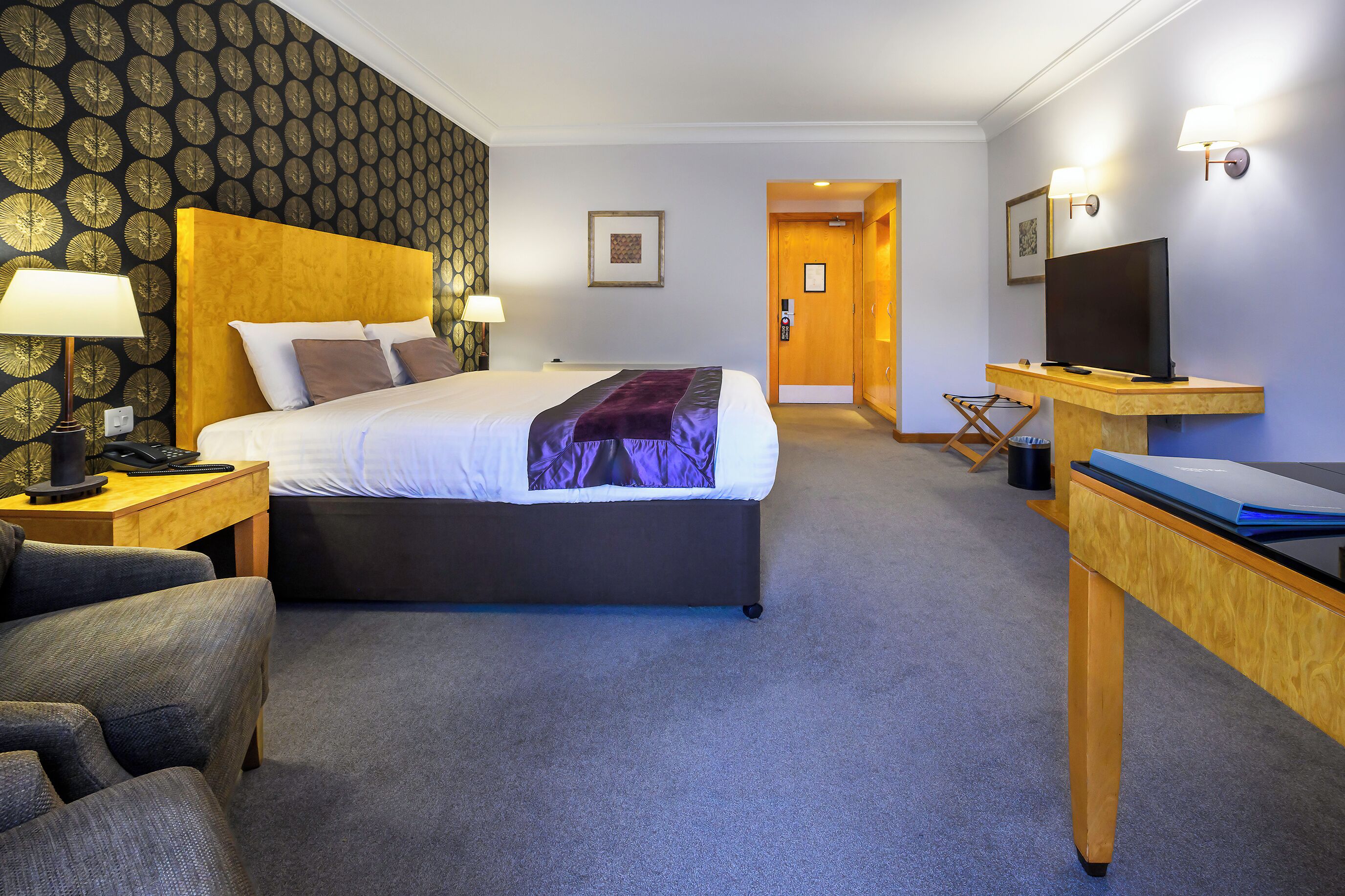 A hotel room with a bed, a desk with a computer, a chair, a TV, and a lamp.