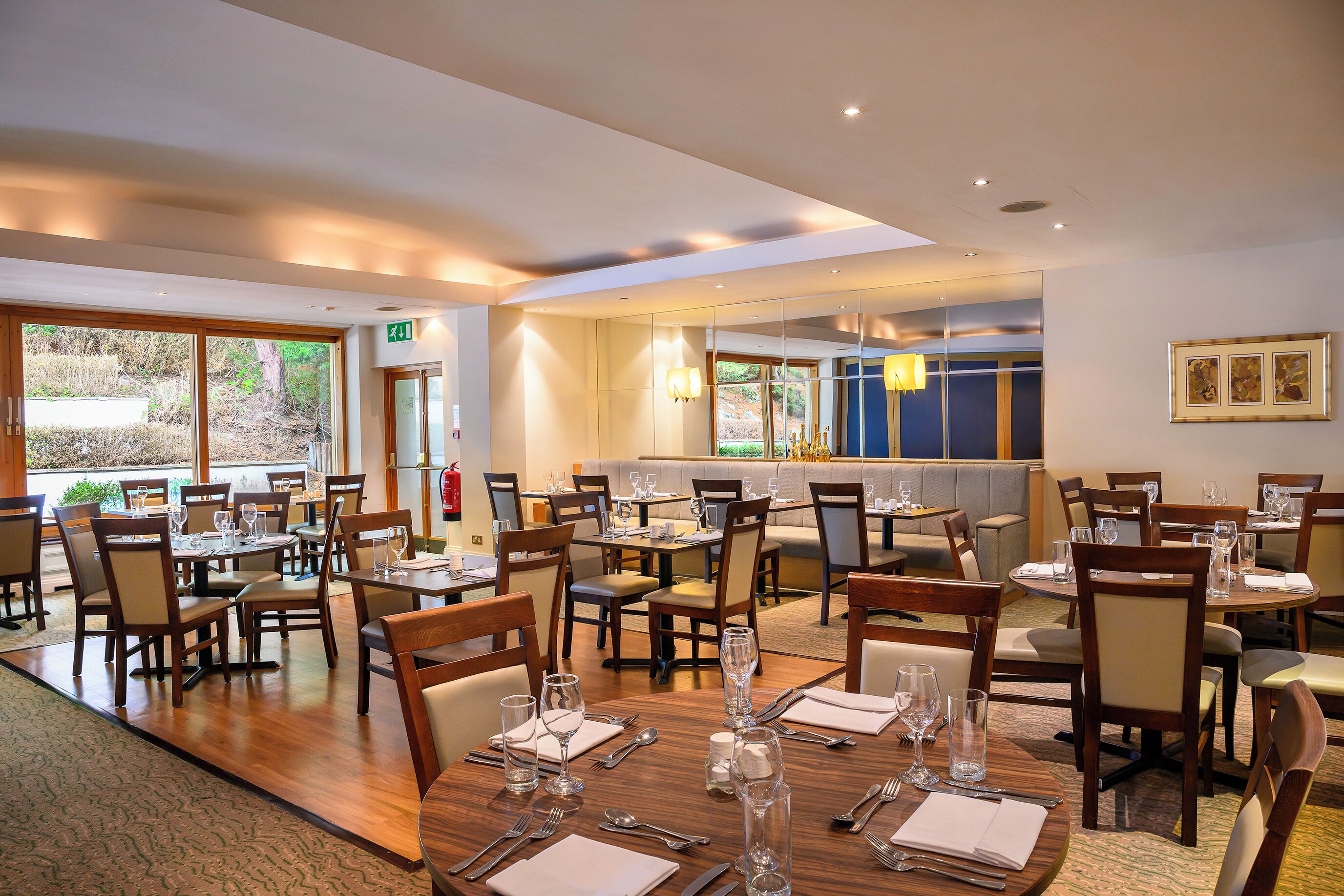 A restaurant with multiple dining tables and chairs, a bar area, and large windows offering a view of the outdoors.
