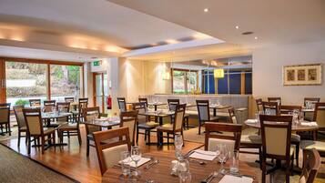 A restaurant with multiple dining tables and chairs, a bar area, and large windows offering a view of the outdoors.