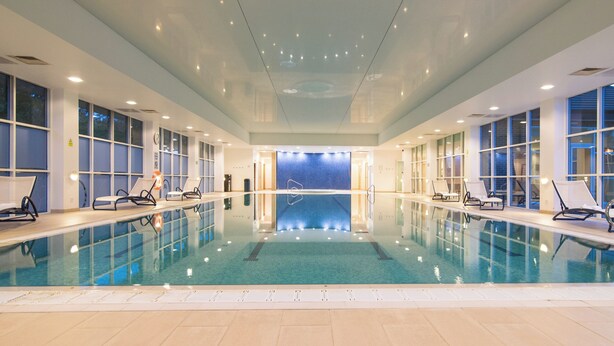 An indoor swimming pool with clear water, surrounded by lounge chairs and large windows. - Regency Park Hotel (Thatcham)