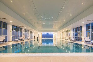 An indoor swimming pool with clear water, surrounded by lounge chairs and large windows. - Regency Park Hotel (Thatcham)