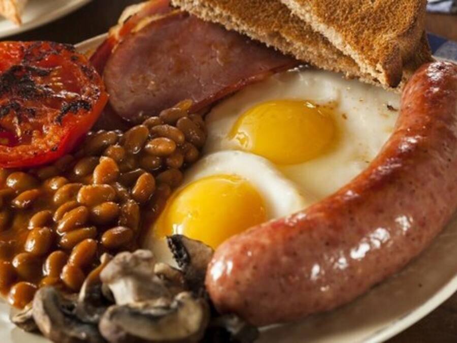 Een bord met een gegrilde tomaat, bonen, een gebakken ei, een plak ham, een worst en een sneetje toast.