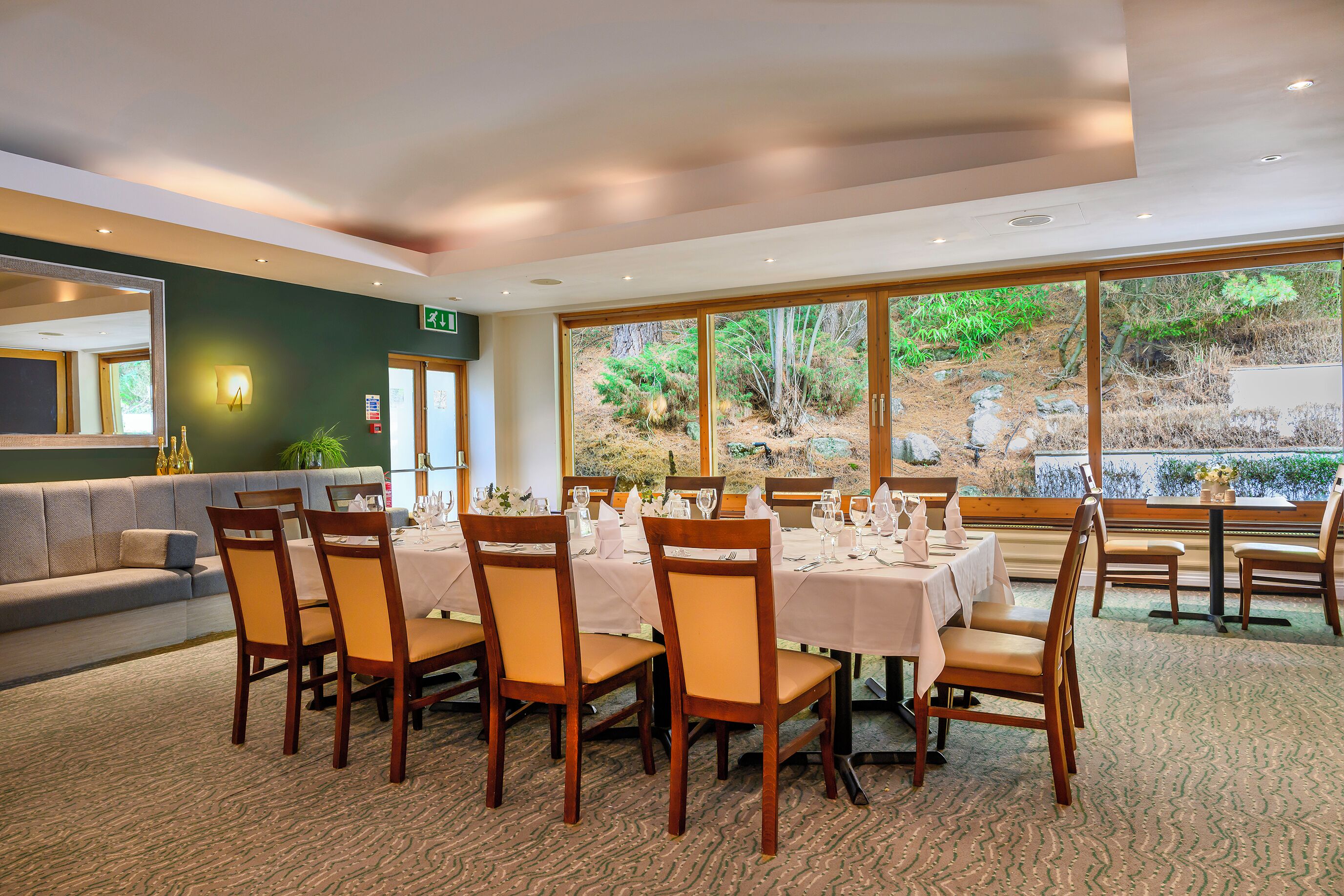 Uma sala de jantar com mesas preparadas para seis pessoas, cadeiras de madeira e vista para uma paisagem natural através de grandes janelas.