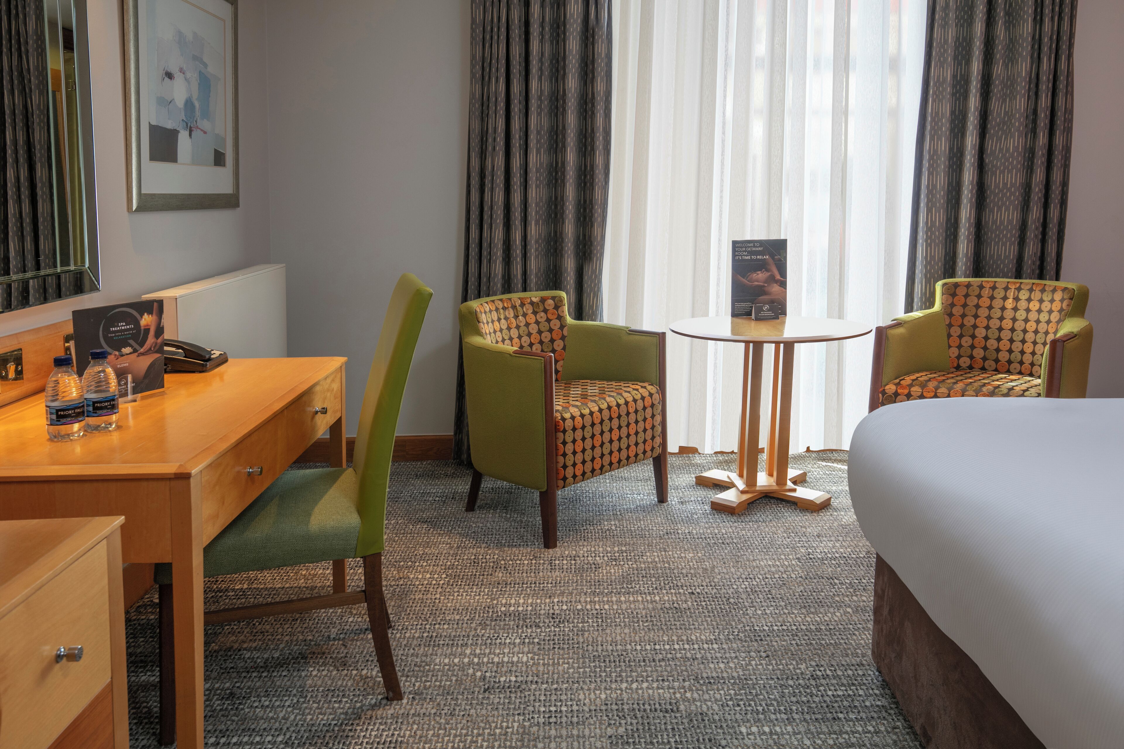 Habitación de hotel con cama, dos sillones verdes estampados, una mesita redonda, un escritorio con teléfono y un cuadro en la pared.